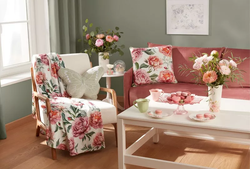 Przytulny salon z produktami Pepco: różowa sofa, fotel z kwiatowym kocem, poduszka motyl i świeże kwiaty w wazonach.
