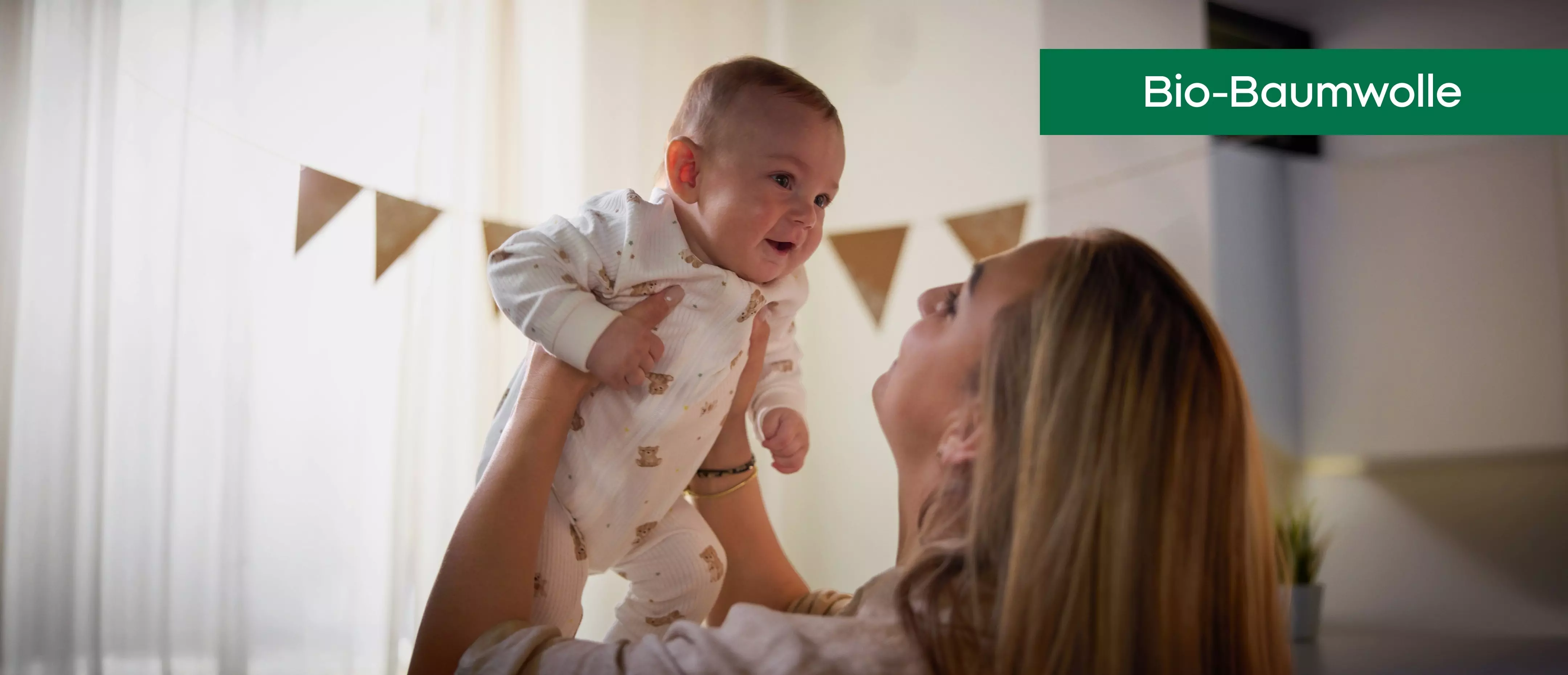 Pepco Bio-Baumwolle Babykleidung, Mutter hebt lächelndes Baby in gemustertem Strampler hoch.