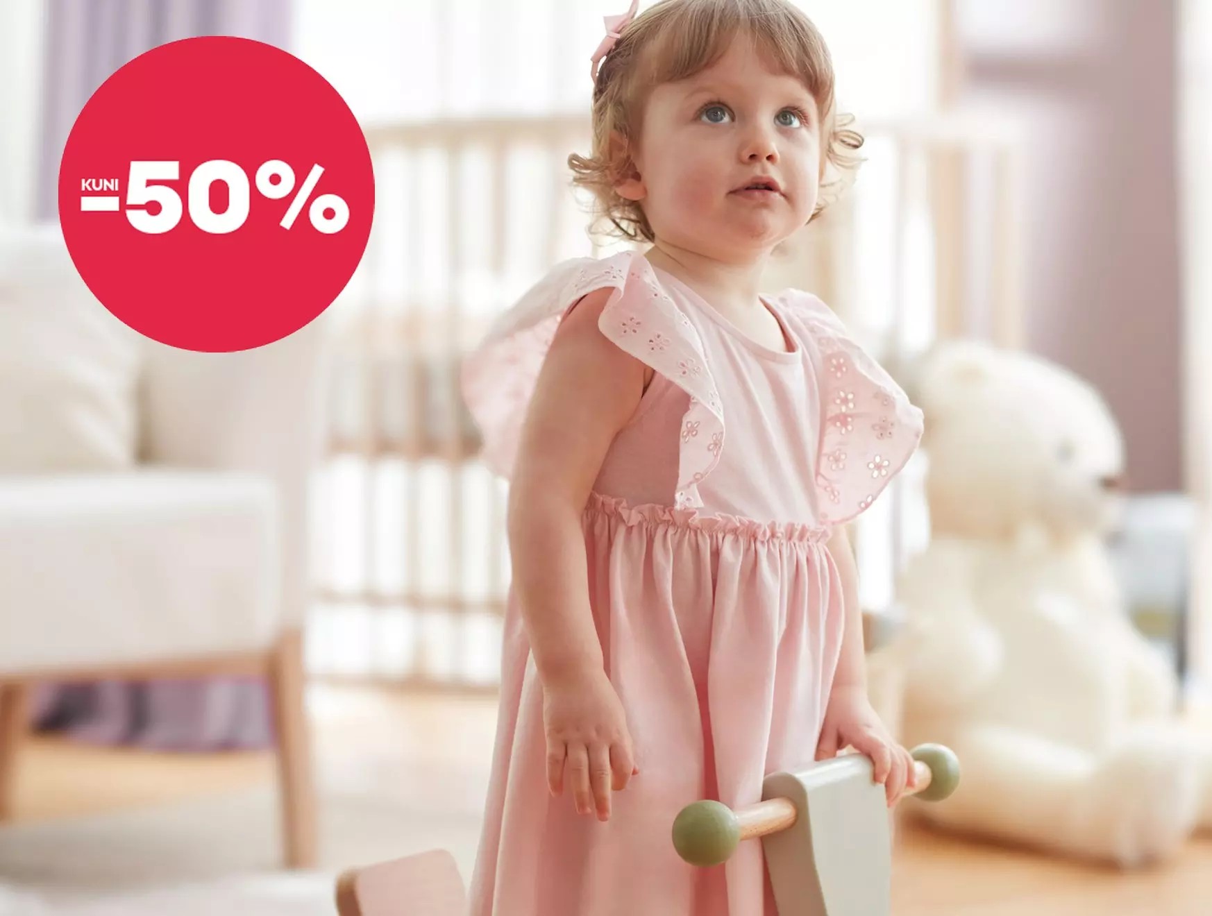 Pepco toddler wearing a pink dress with floral embroidery, holding a wooden toy with KUNI -50% offer badge.