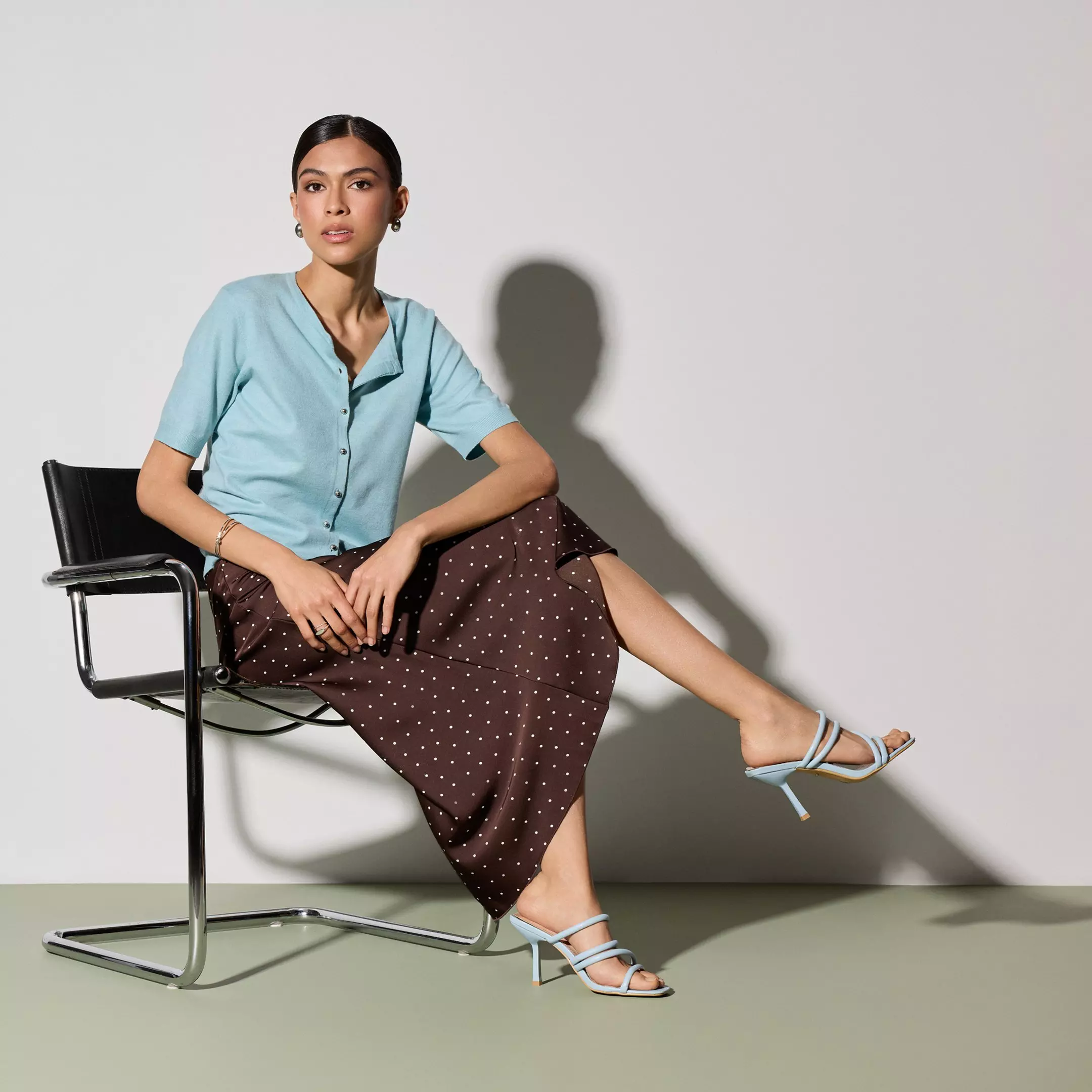 Mujer sentada con camisa azul Pepco y falda marrón de lunares blancos, combinada con sandalias azules.