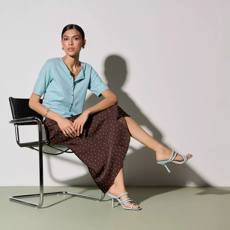 Mujer sentada con camisa azul Pepco y falda marrón de lunares blancos, combinada con sandalias azules.
