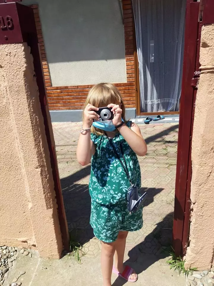 Copil fotografiind cu un aparat foto instant, purtând o rochie verde cu imprimeu floral.