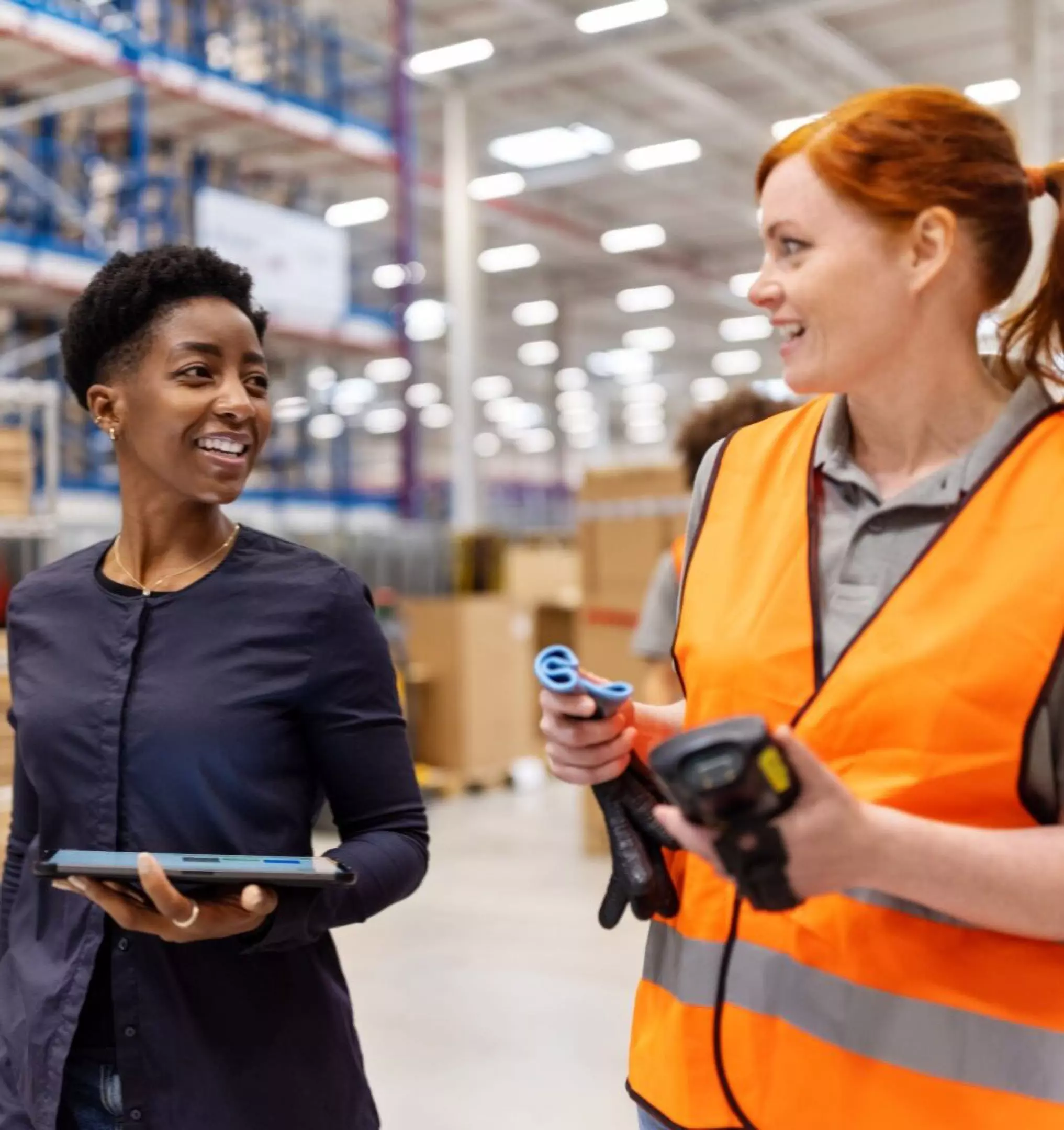 Two colleagues conversing in a Pepco warehouse, one holding a tablet while the other in safety vest holds scanning equipment.