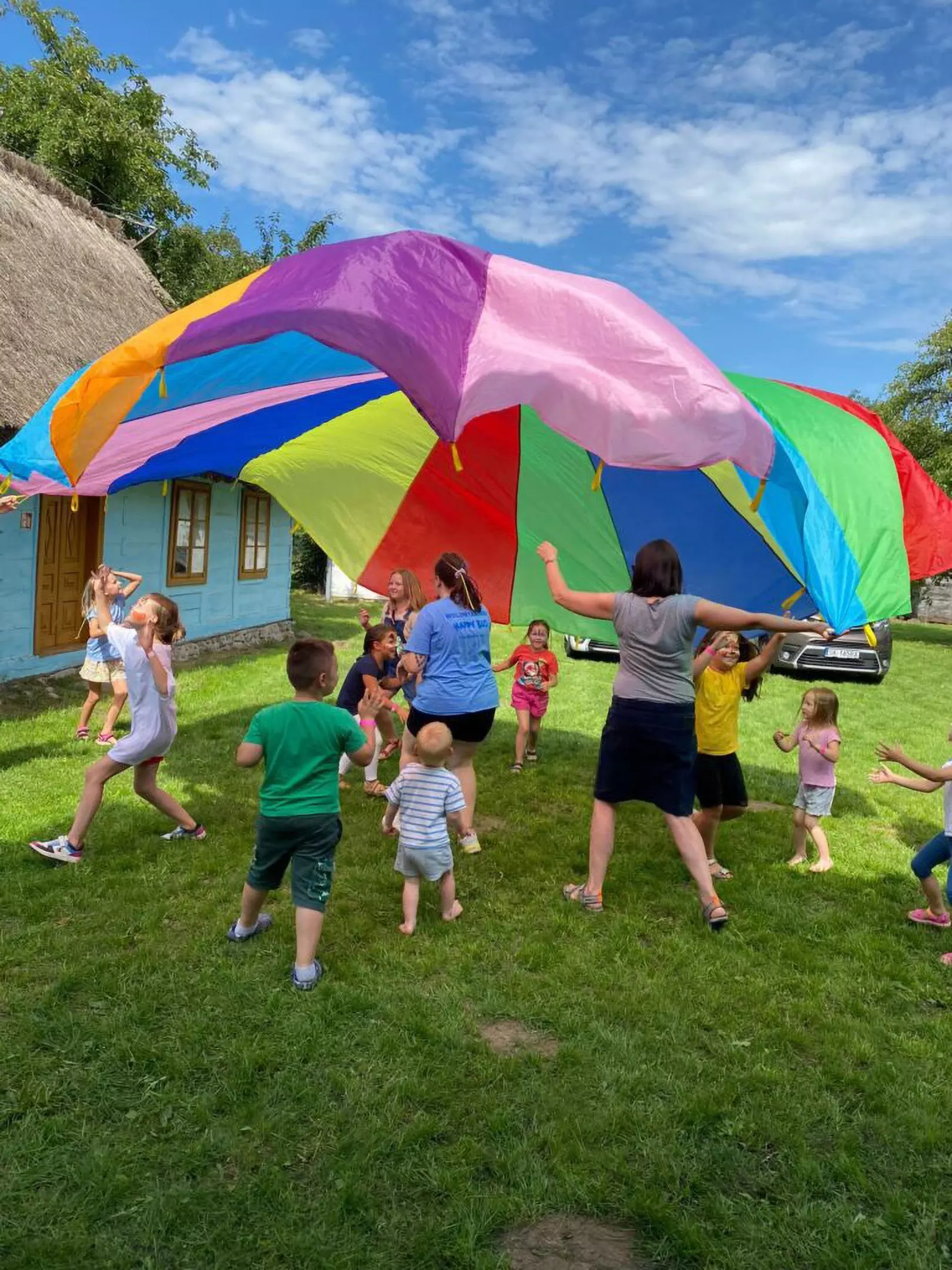 Children playing with a colorful Pepco parachute outdoors on grass, with adults guiding the activity near a blue cottage.