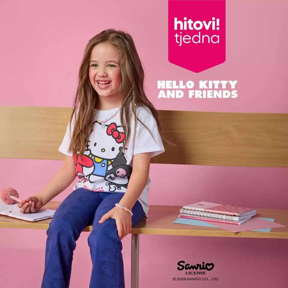 Pepco djevojčica u Hello Kitty majici i plavim hlačama sjedi na stolu s bilježnicama i osmjehuje se.