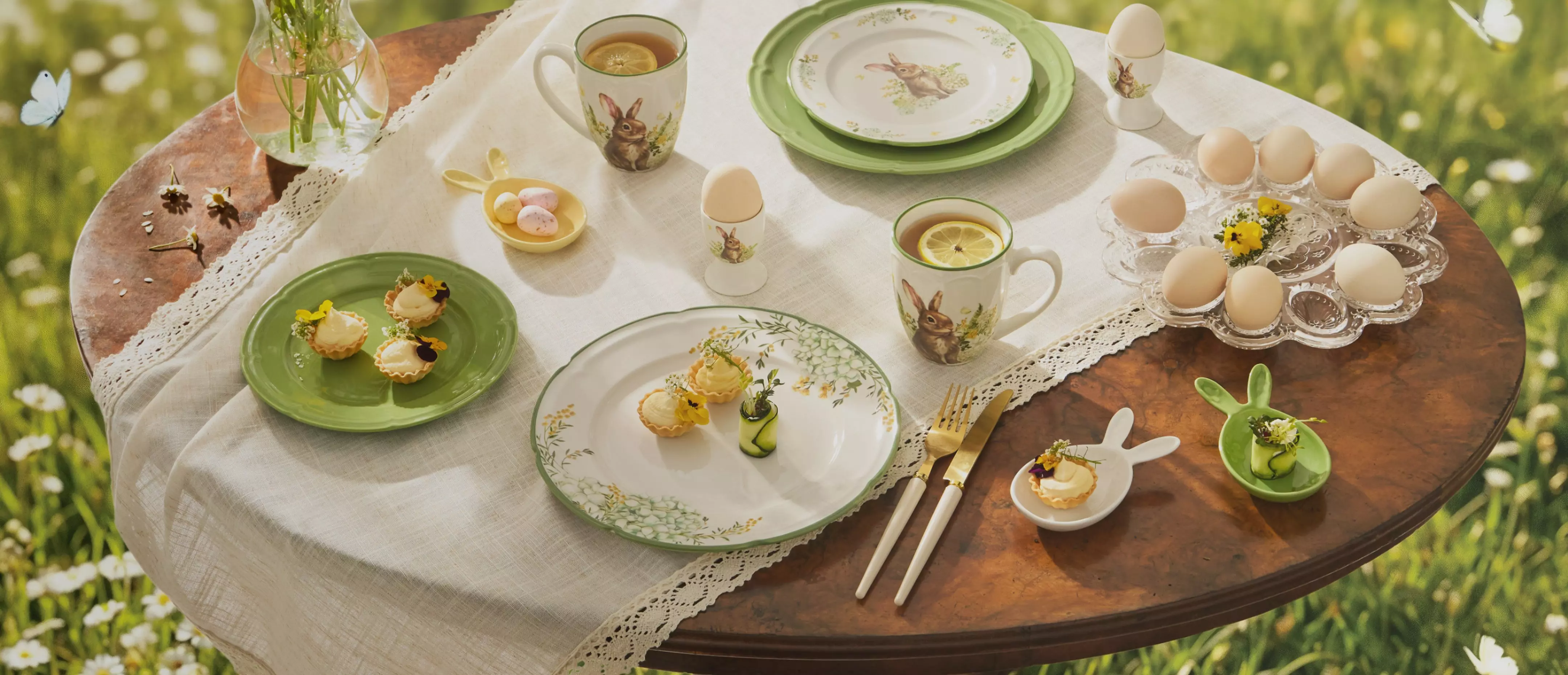 Wielkanocne nakrycie stołu z zastawą ozdobioną motywami królików, jajkami, herbatą z cytryną i małymi tarteletkami na łące.