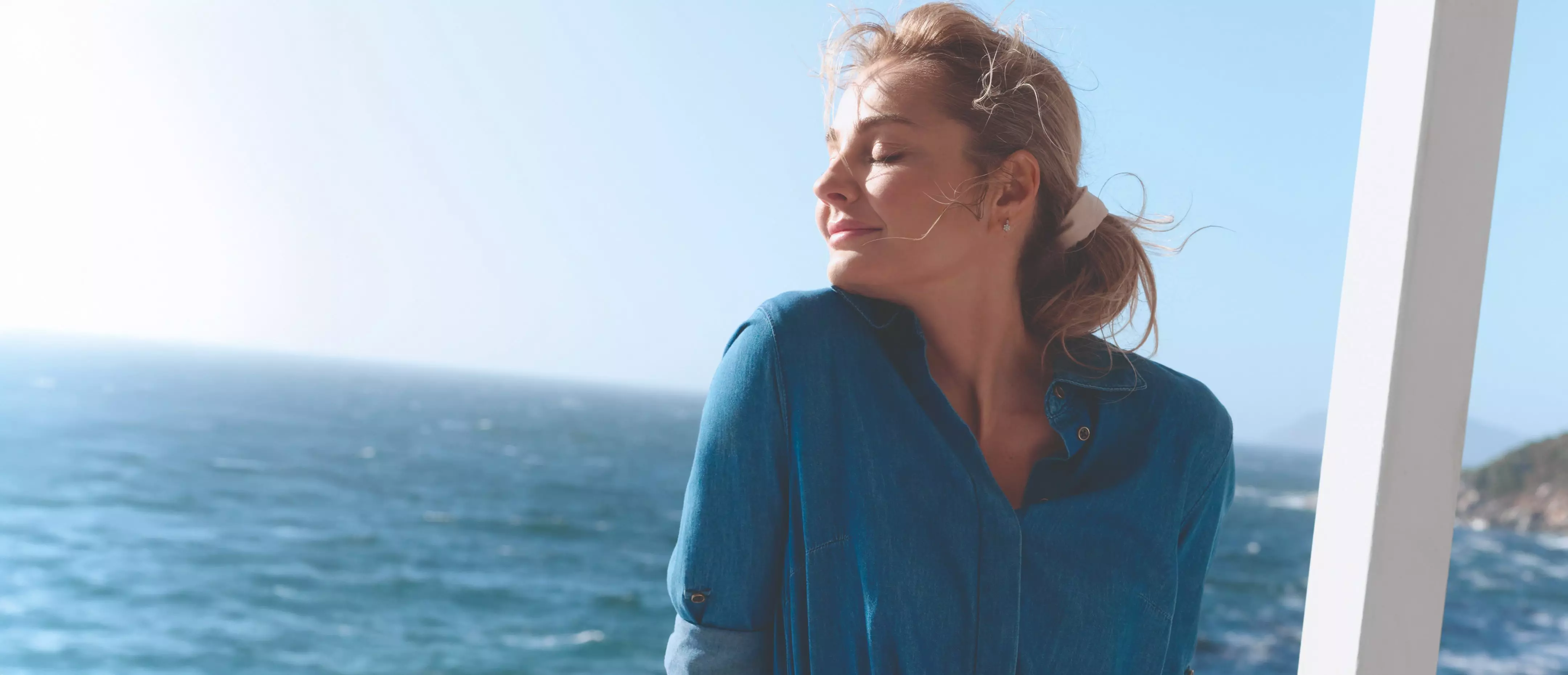 Kobieta w niebieskiej bluzce cieszy się słonecznym dniem nad morzem z rozwianymi włosami.