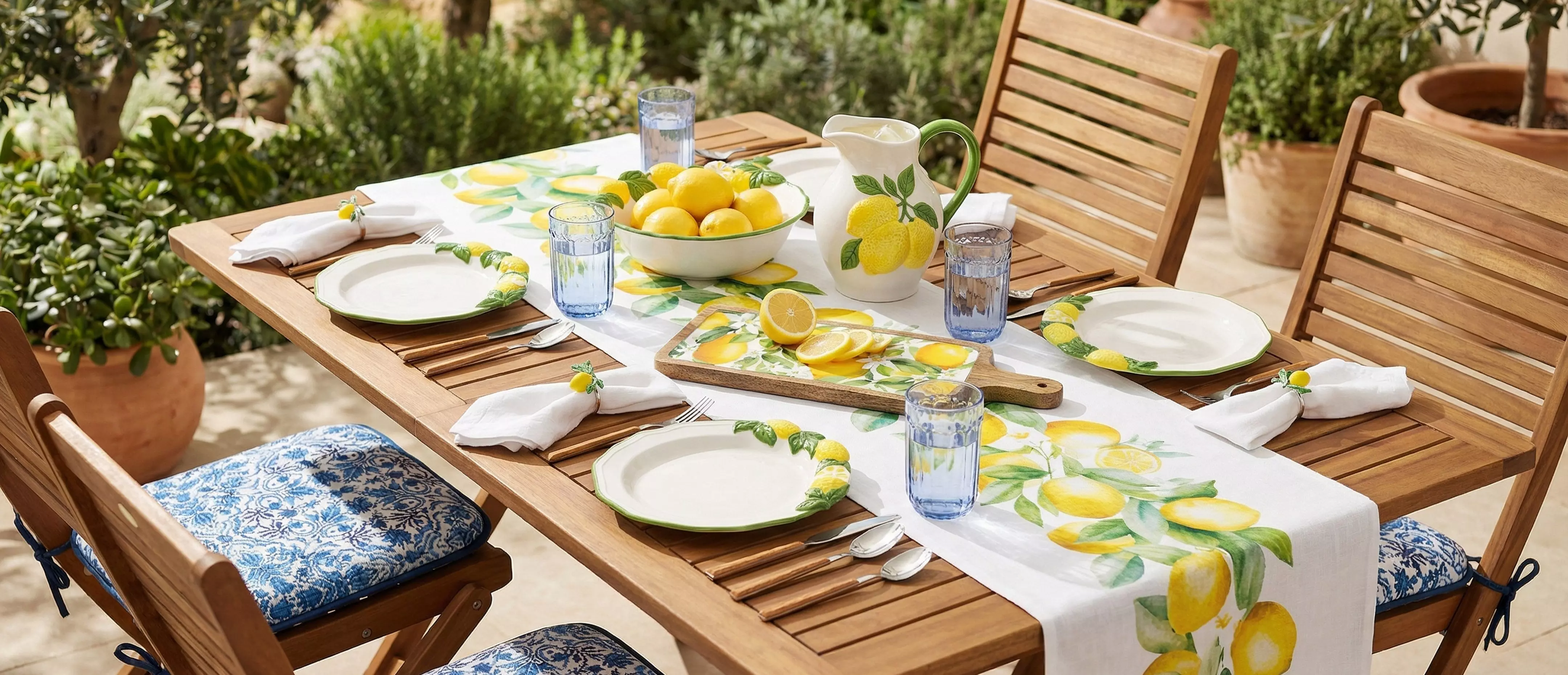 Dřevěný zahradní stůl prostřený s citronovým motivem, talíři, sklenicemi a mísou citronů.