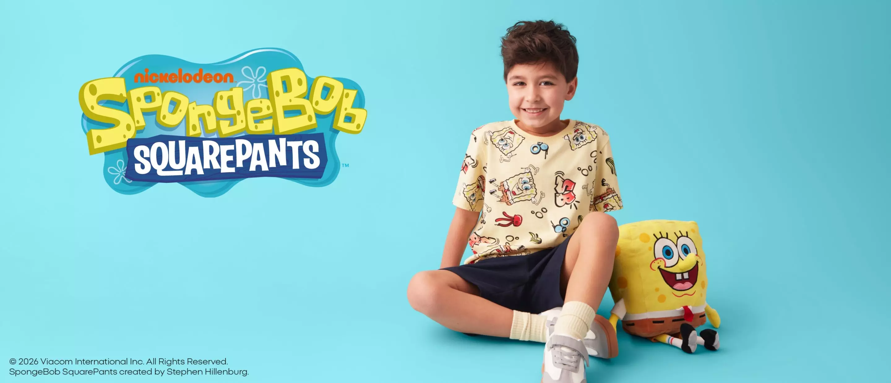 Chłopiec w beżowej koszulce z motywem SpongeBob siedzi obok pluszowej zabawki SpongeBob SquarePants.