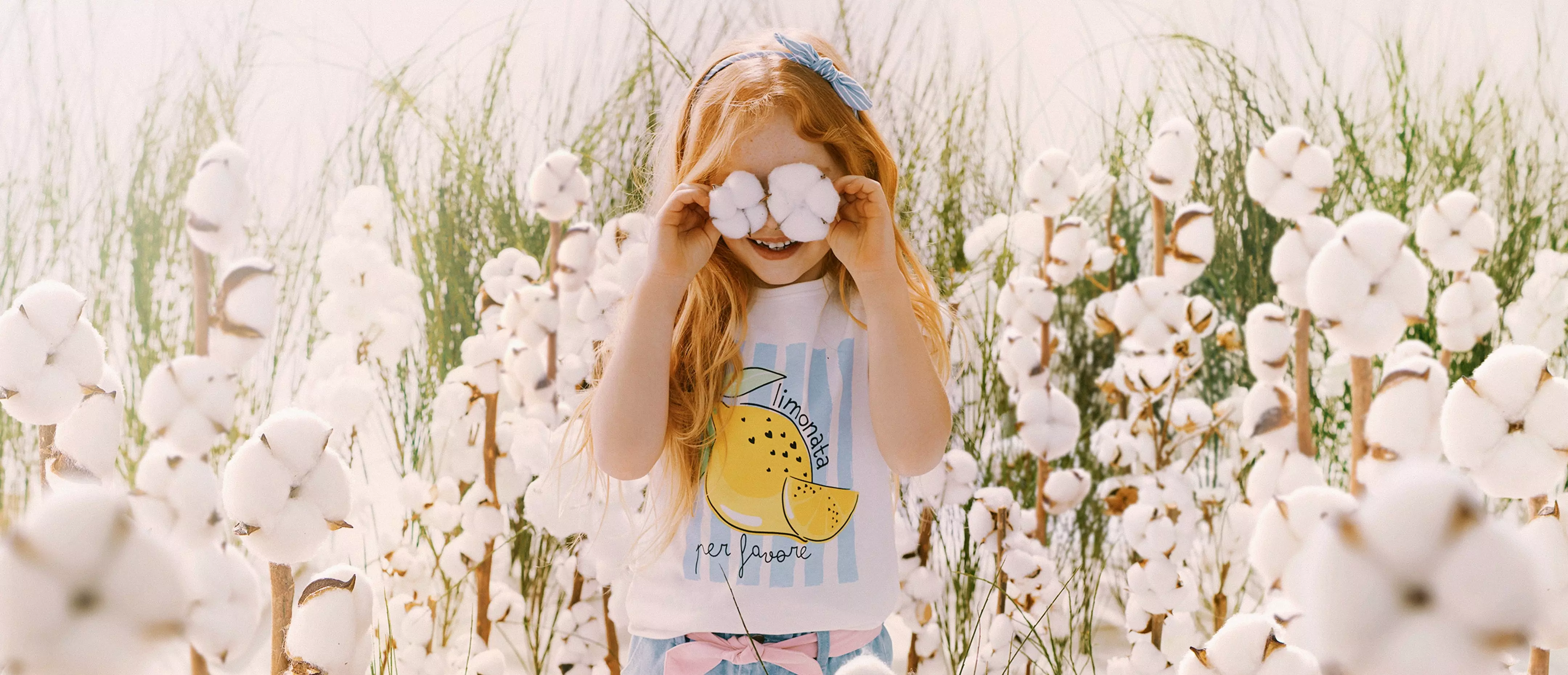 Niña pelirroja sosteniendo flores de algodón sobre sus ojos en un campo de algodón, viste camiseta con limones.