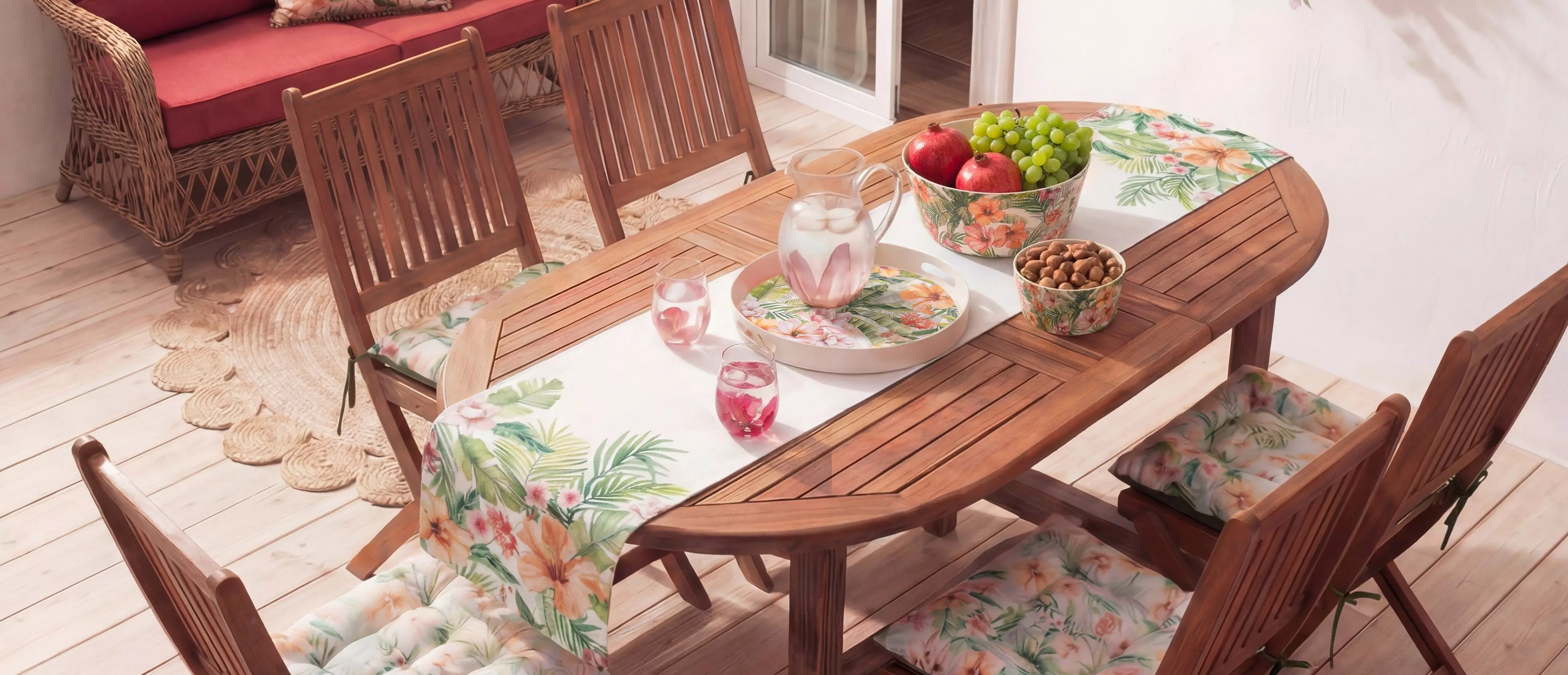 Mesa de comedor de madera ovalada con sillas y cojines estampados tropicales, servida con frutas y bebidas.