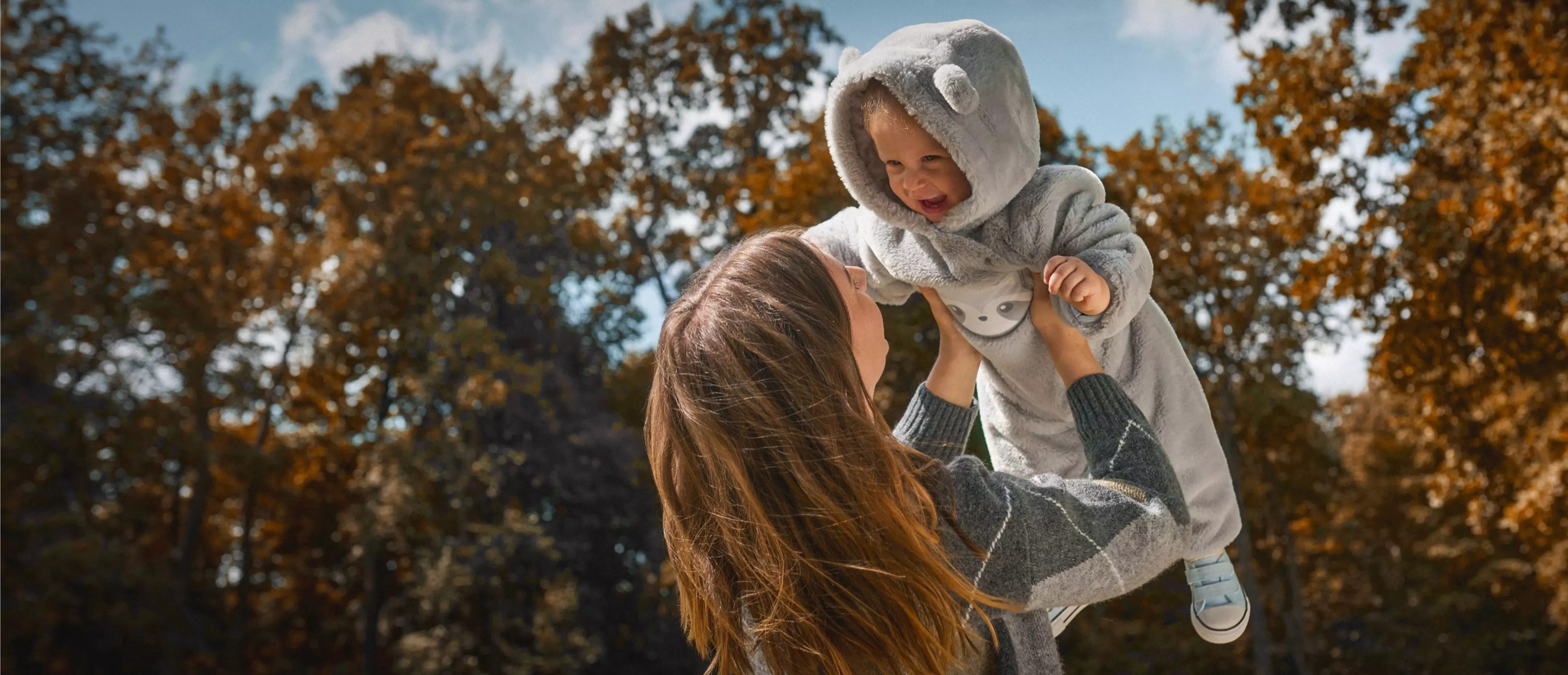 Mamma solleva gioiosamente bambino che indossa tuta con cappuccio grigia Pepco all'aperto in autunno.