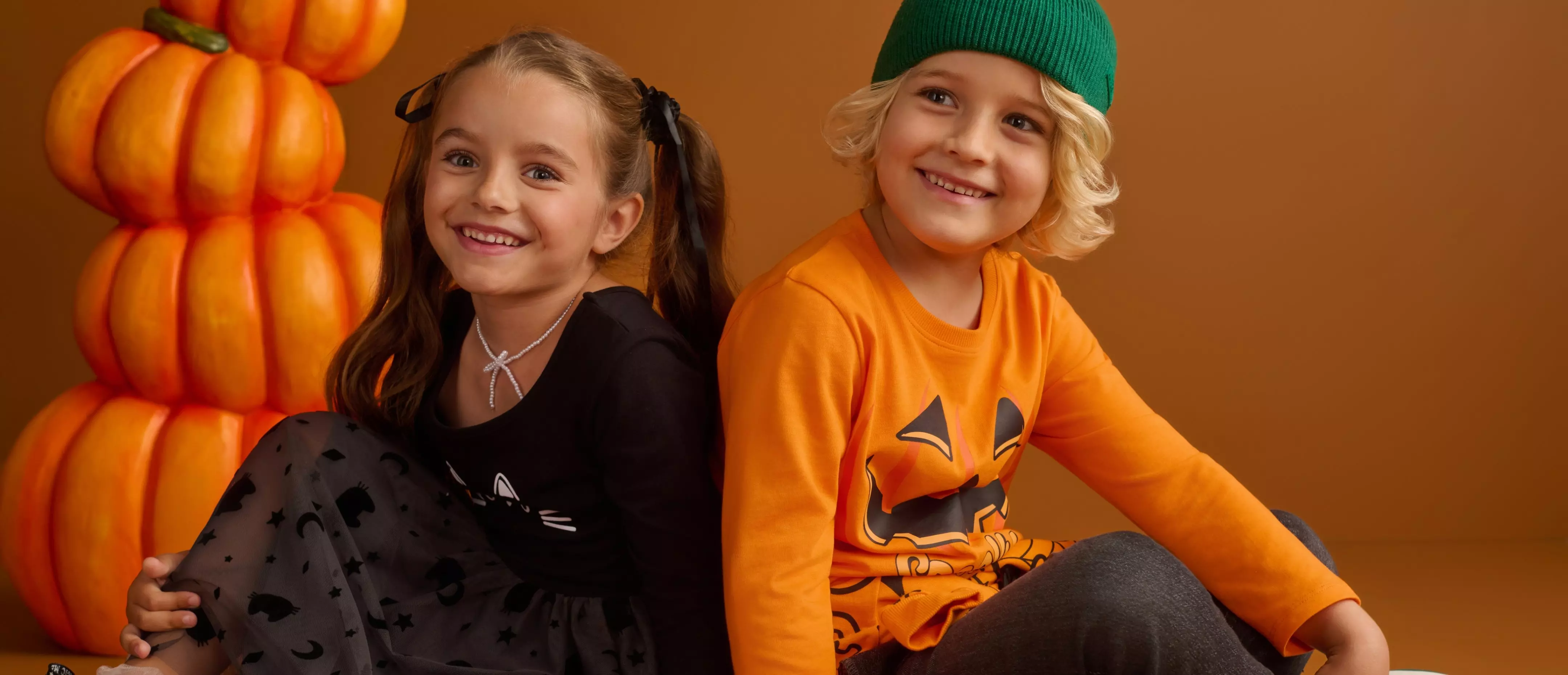 Dve usmiате deti v halloweenskom oblečení Pepco sedia pred oranžovými tekvicami.