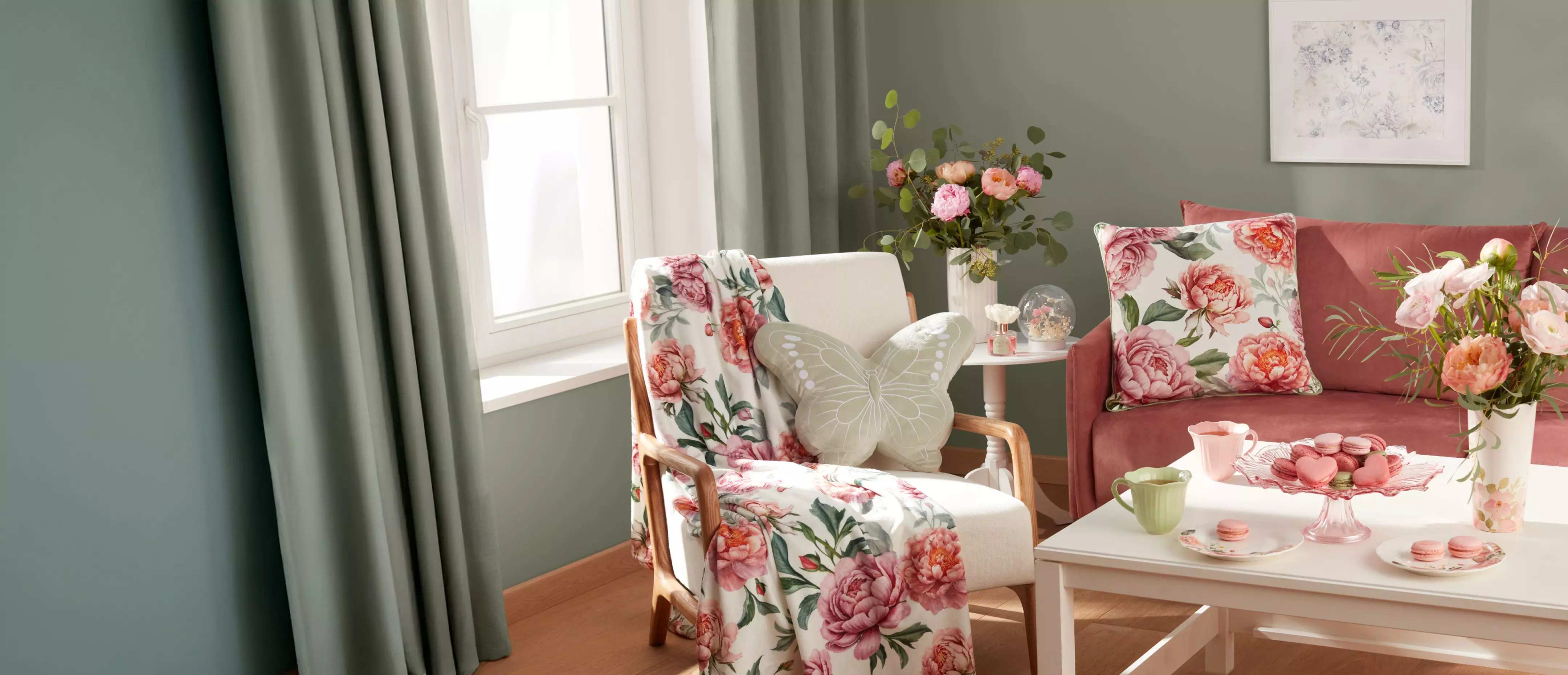 Przytulny salon z meblami Pepco, fotel z kwiatowym kocem, różowa sofa z poduszkami i stolik z makaronikami.