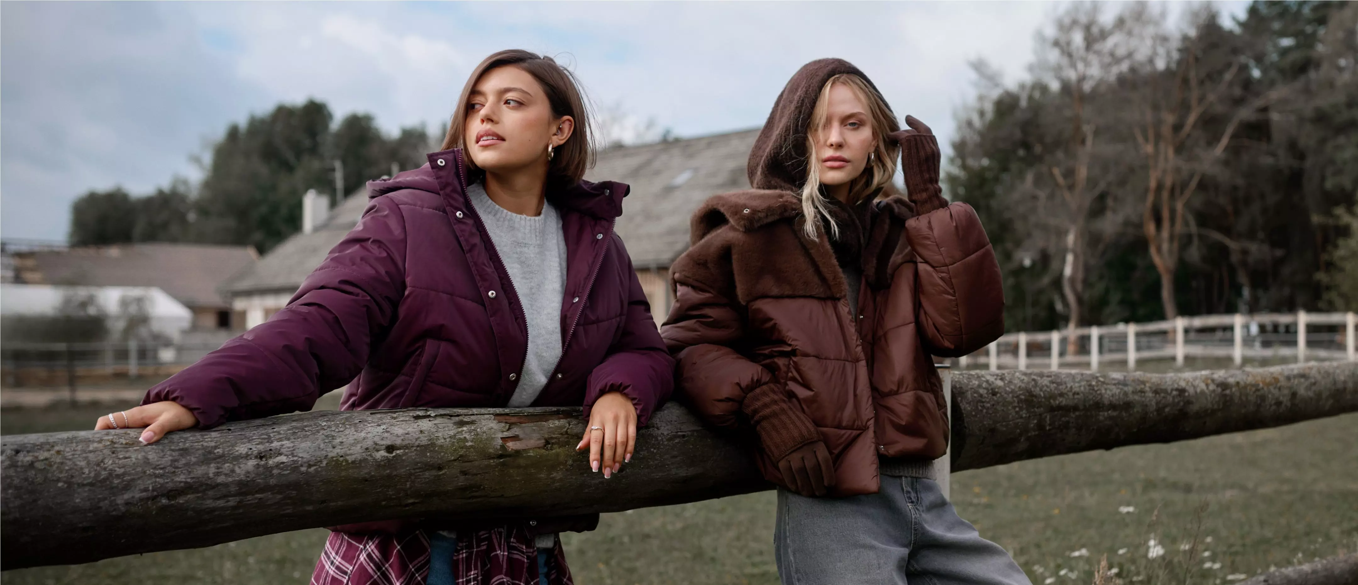 Two women wearing Pepco puffer jackets in burgundy and brown leaning on a wooden fence outdoors.