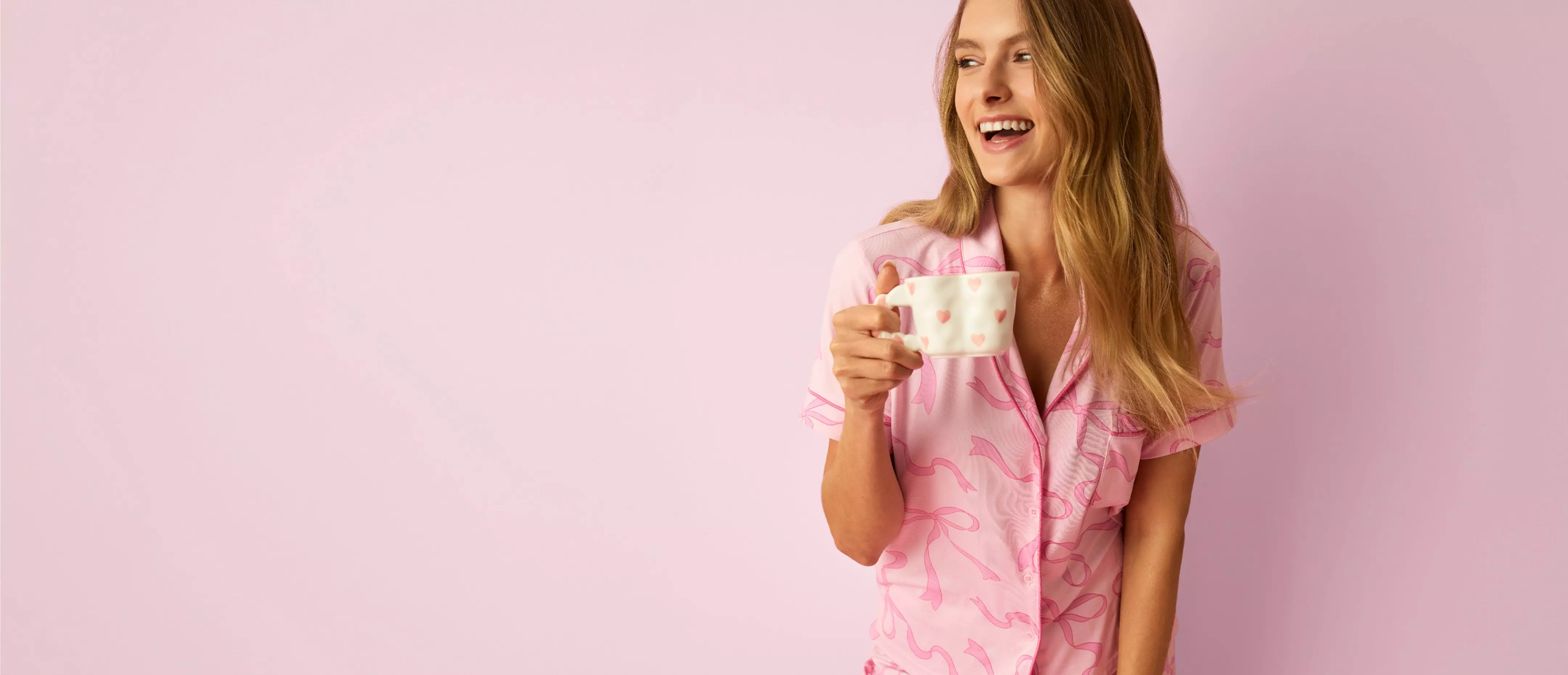 Osoba w różowej piżamie Pepco trzymająca biały kubek z różowymi serduszkami, uśmiechająca się na różowym tle.