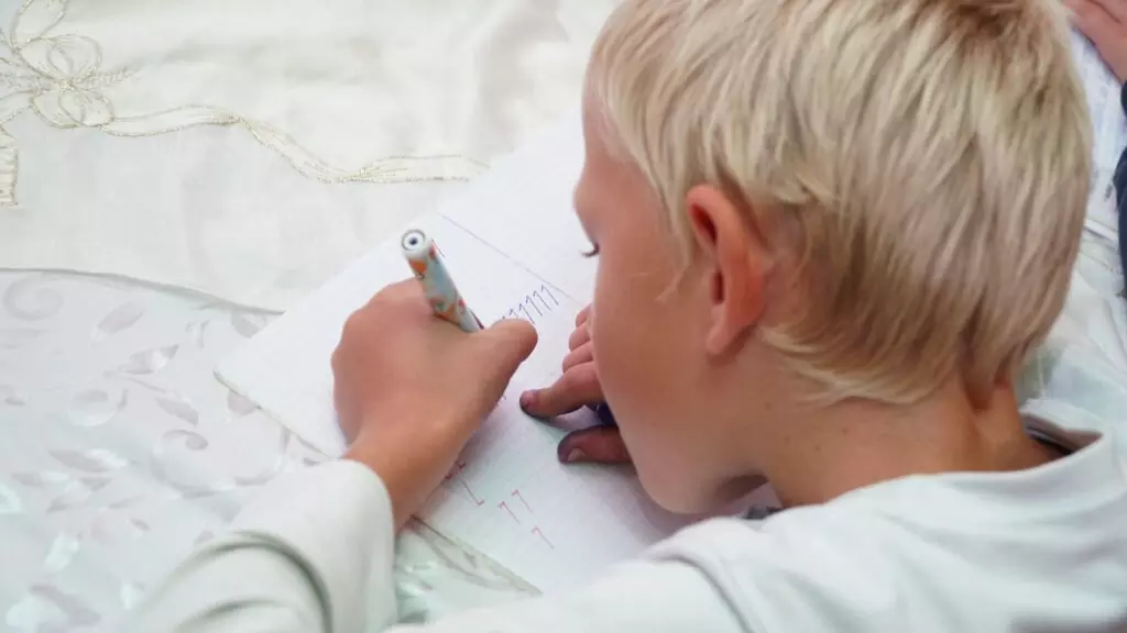 Copil cu păr blond desenând cu un creion colorat într-un caiet cu linii pătrate.