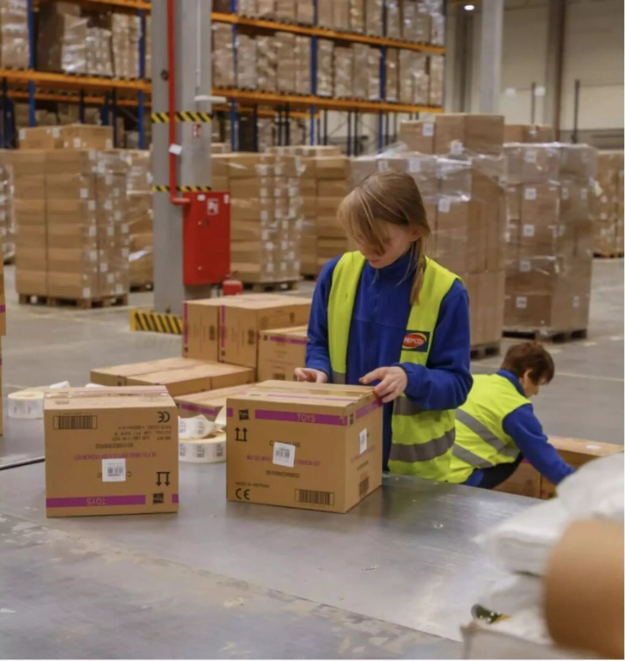 Pepco warehouse workers in safety vests processing toy boxes, surrounded by stacked inventory pallets.