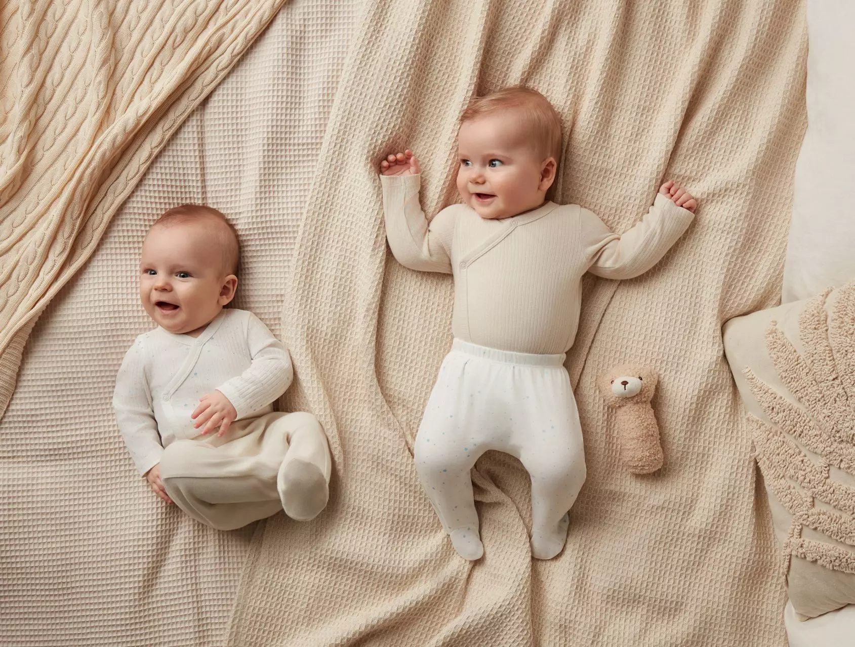 Dwoje niemowląt leży na beżowych kocach, ubrane w kremowe ubranka z dzianiny.