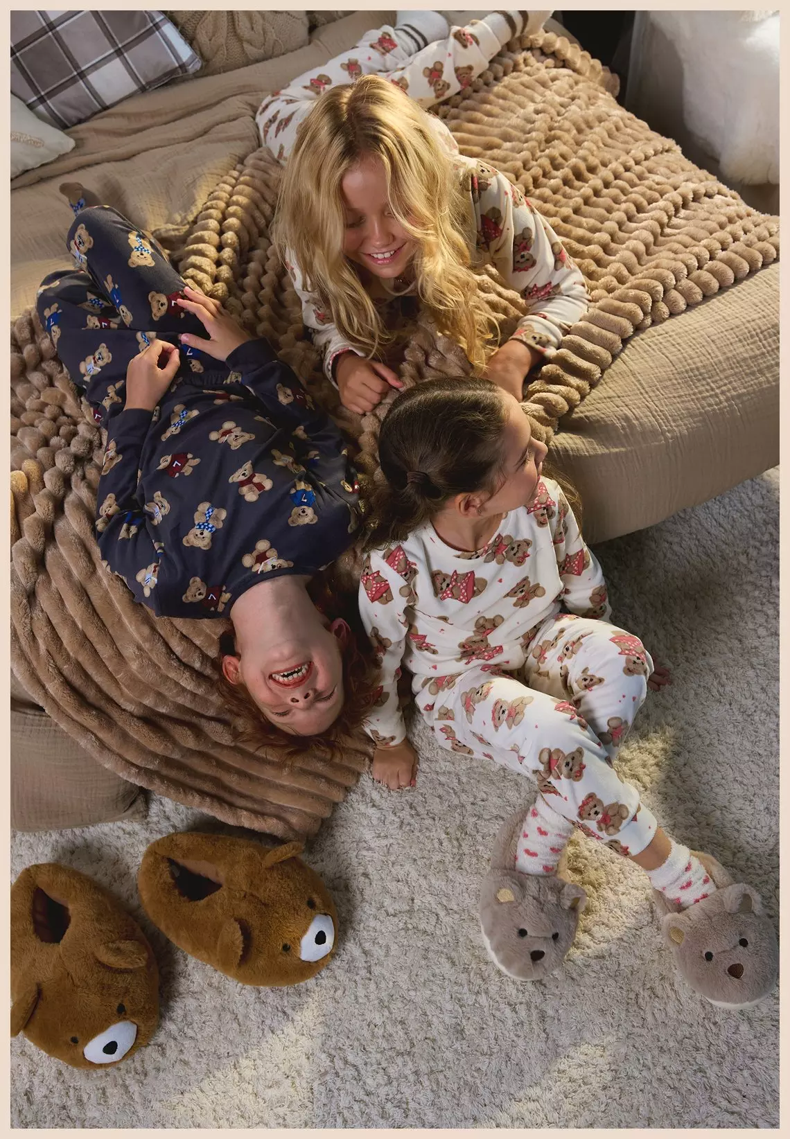 Tres niños riendo juntos en pijamas estampados de ositos de Pepco sobre una manta tejida beige.