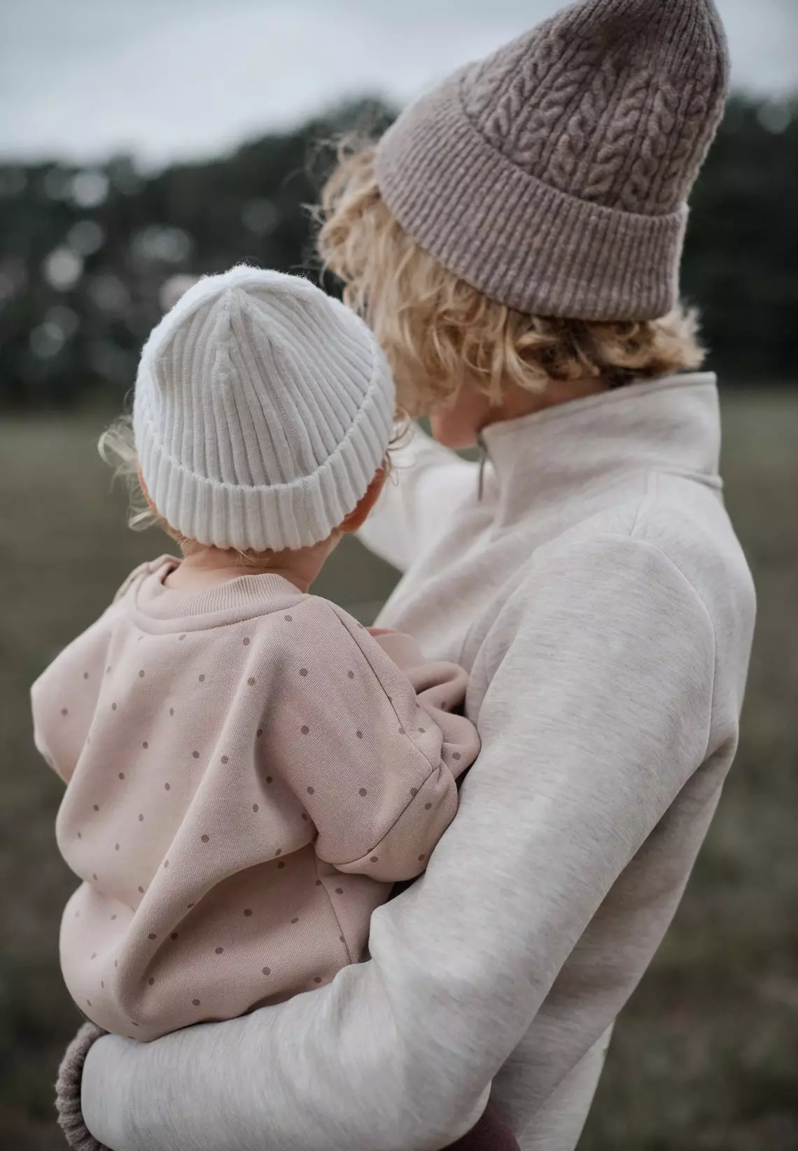 Adulto llevando a un niño, ambos vistiendo gorros de punto Pepco en tonos neutros al aire libre.