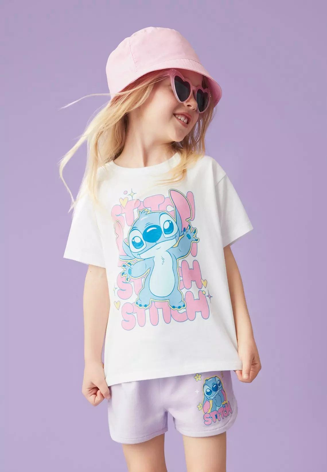 Child wearing white Stitch graphic t-shirt, pink bucket hat and heart-shaped sunglasses, smiling.