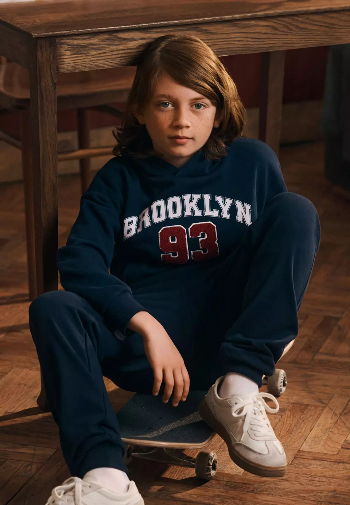 Băiat purtând hanorac Pepco albastru cu imprimeu BROOKLYN 93 și pantaloni bleumarin, așezat pe skateboard.