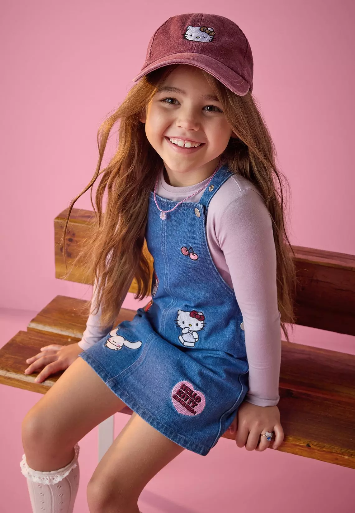 Fată zâmbitoare purtând sarafan din denim Hello Kitty și șapcă Pepco, șezând pe o bancă de lemn.