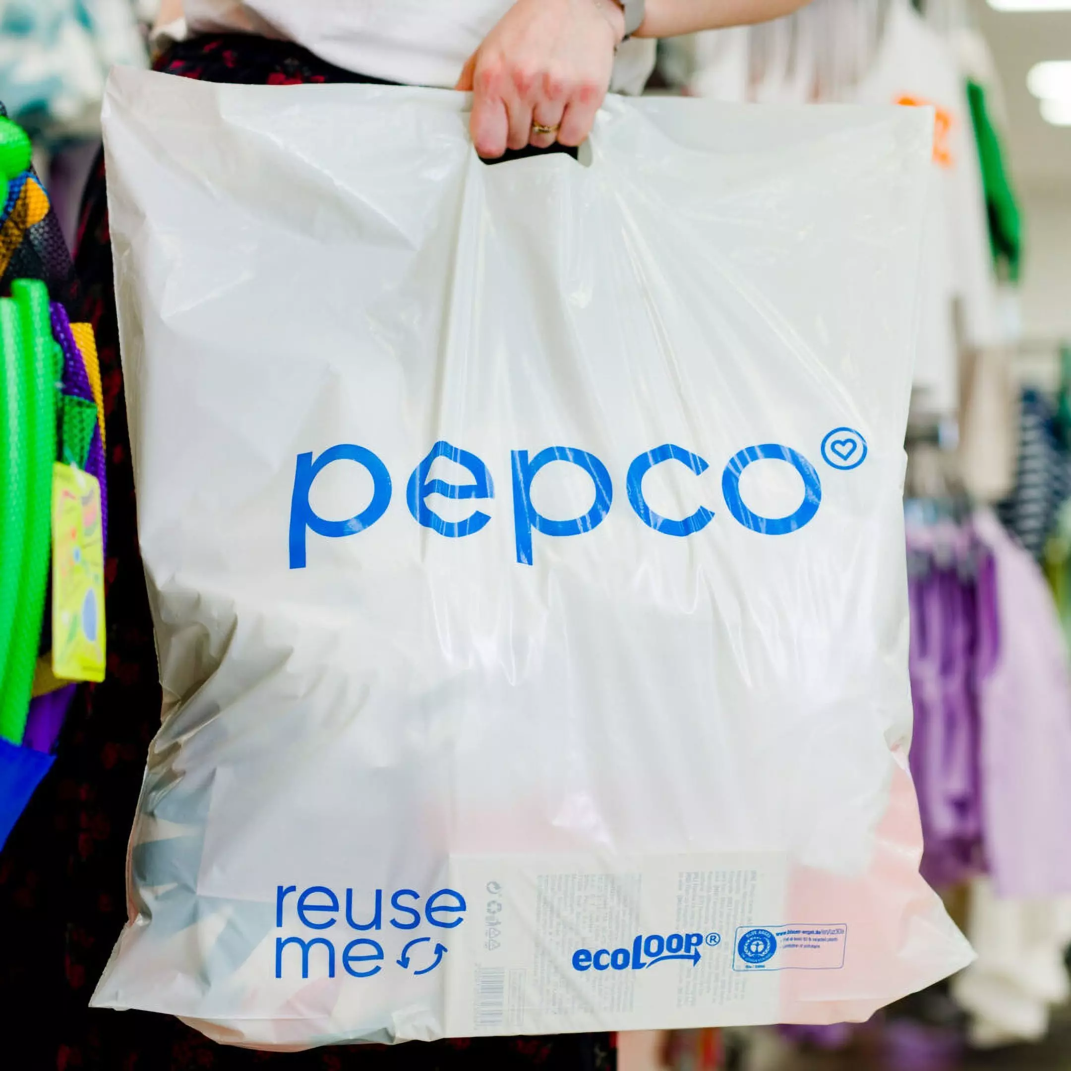 Person holding a white Pepco shopping bag with blue logo and "reuse me" text in a retail store environment.