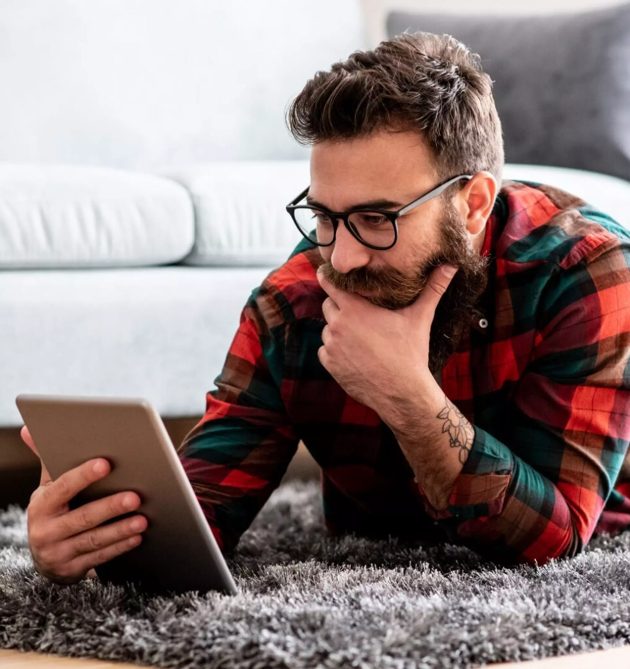 Person in Pepco plaid shirt lying on a fluffy rug, thoughtfully browsing a tablet while wearing glasses.