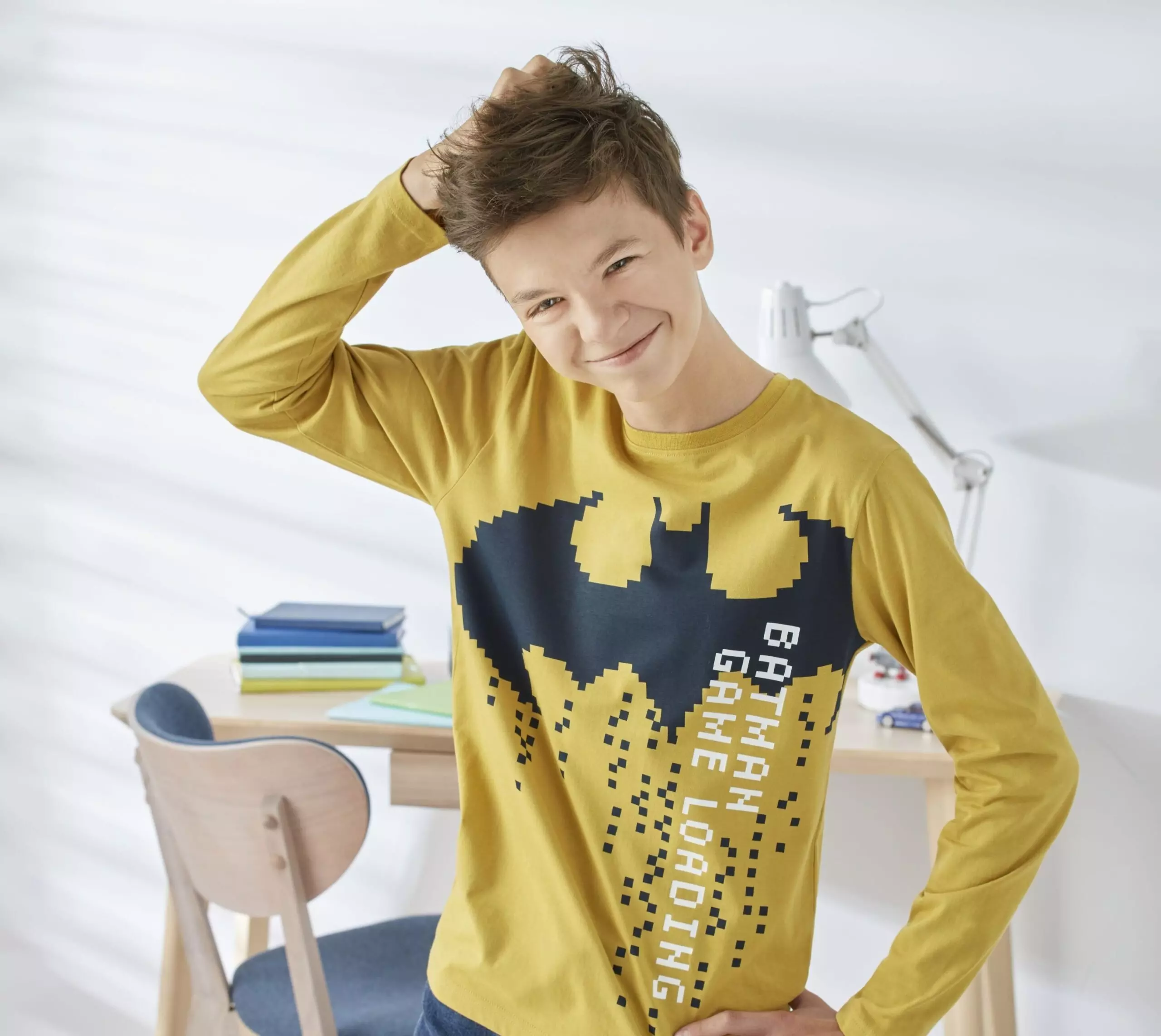 Młoda osoba w żółtej bluzce Pepco z grafiką Batmana i napisem "Gotham Game Loading", uśmiechnięta w jasnym pokoju przy biurku.