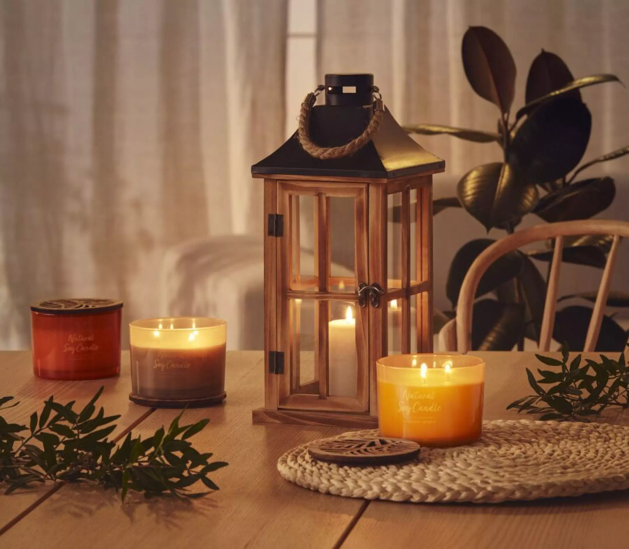 Pepco wooden lantern with lit candle inside, surrounded by scented candles on a wooden table with decorative plants.