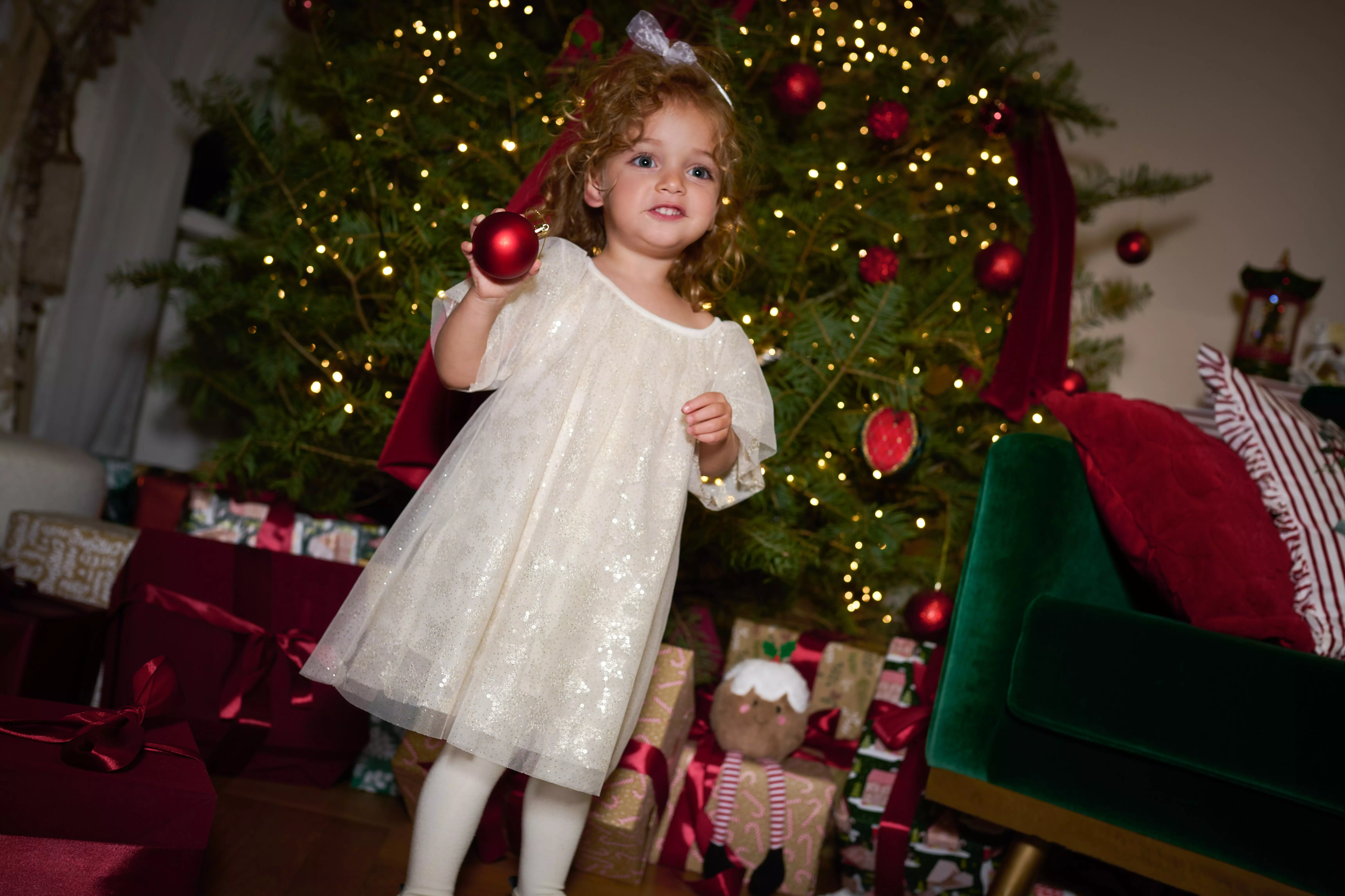 Young girl wearing Pepco sparkly cream dress holding red bauble in front of illuminated Christmas tree.