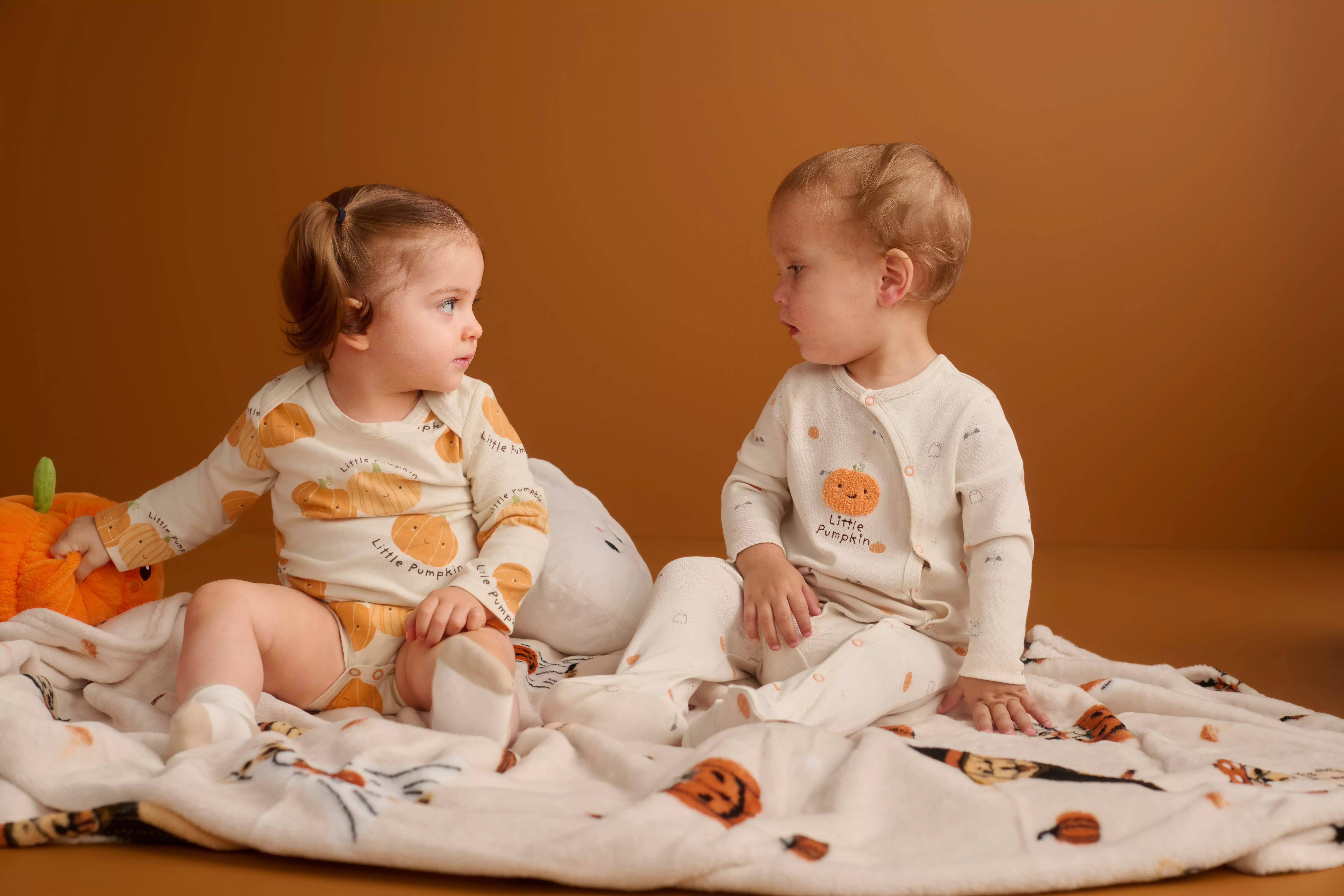 Pepco two babies sitting on a blanket in pumpkin-print sleepsuits, facing each other.