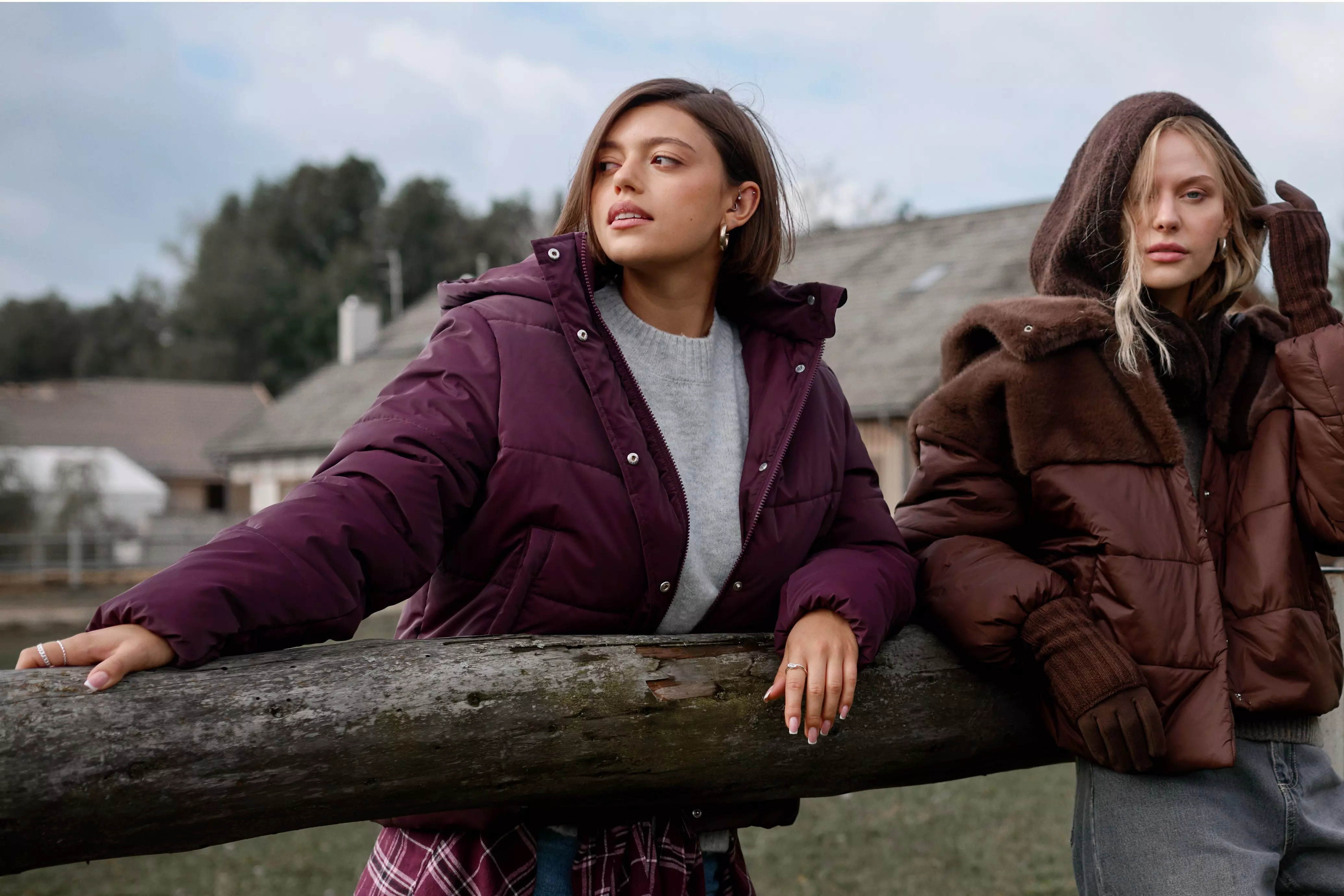 Two women wearing Pepco hooded puffer jackets in burgundy and brown leaning on a wooden fence outdoors.