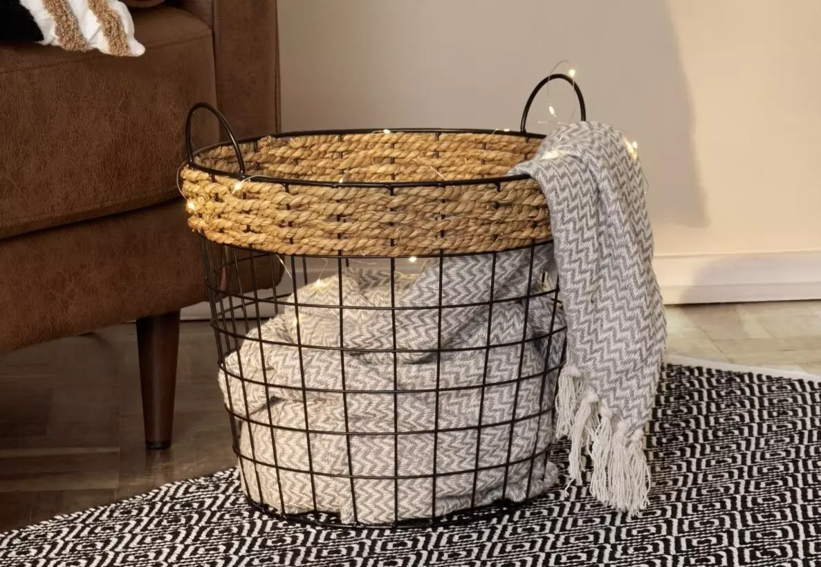 Pepco wire storage basket with woven rattan rim containing folded blankets, placed on patterned rug beside sofa.