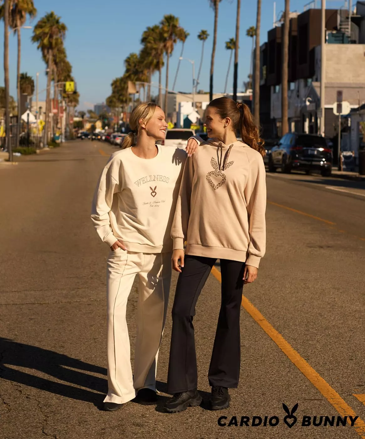 Two women wearing Cardio Bunny sweatshirts walking on a palm tree-lined street. One in cream outfit, other in beige top with black pants.