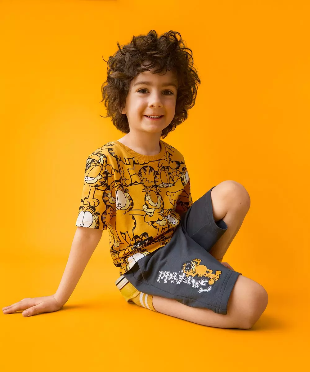 Niño sonriente sentado con camiseta amarilla estampada de Garfield y pantalón corto gris.