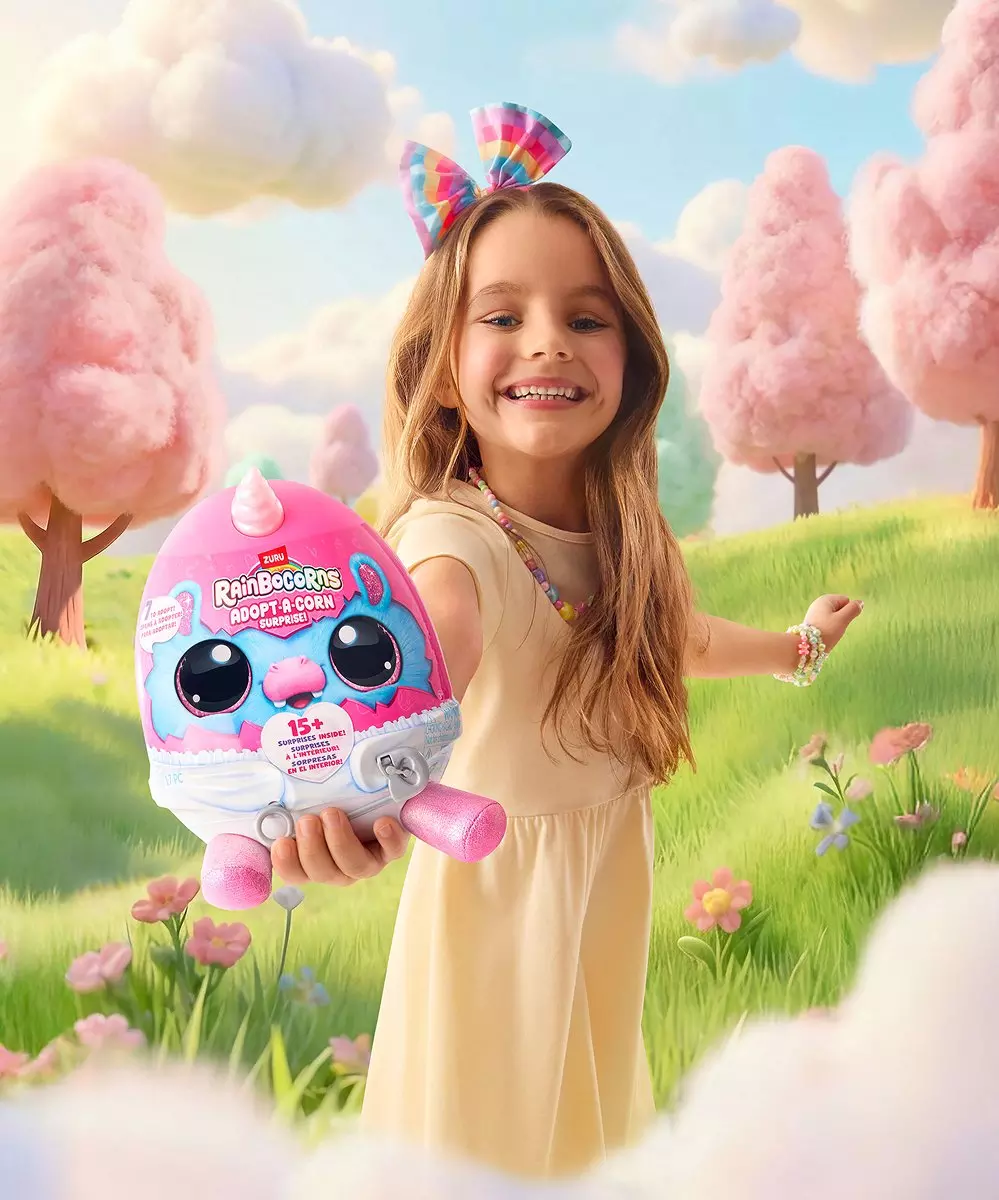 Niña sonriente mostrando juguete Rainbocorns Adopt-A-Corn rosa en un paisaje de fantasía con nubes rosas.