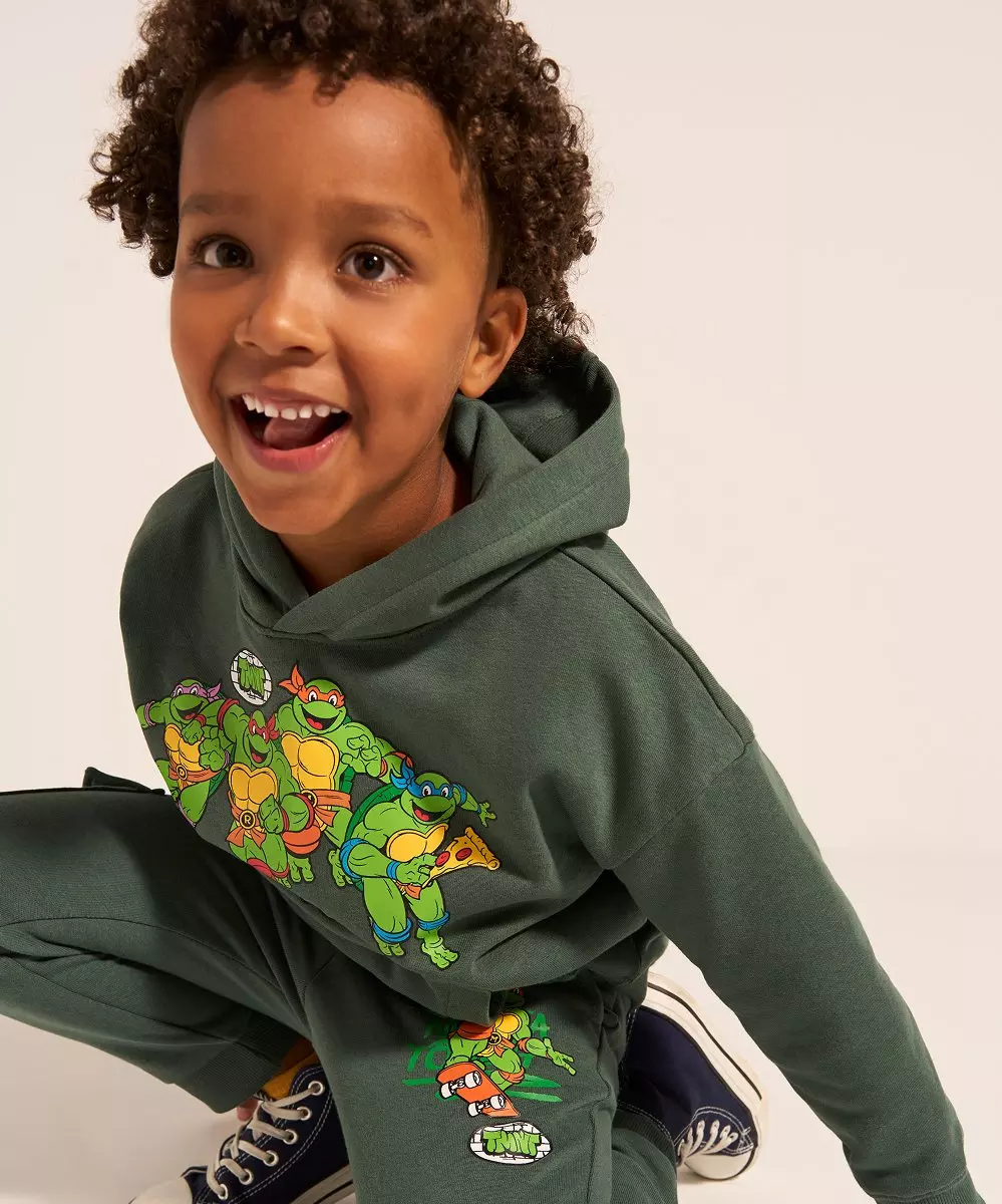 Pepco child wearing green Teenage Mutant Ninja Turtles hoodie and joggers set, smiling playfully.