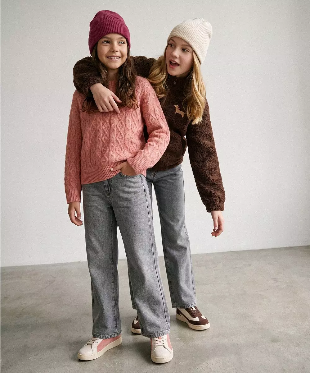 Dos niñas sonrientes de Pepco abrazadas, vistiendo gorros de punto, jerseys y vaqueros grises.