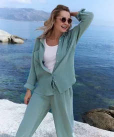 Woman wearing sage green textured co-ord set and sunglasses, posing by the sea with hand in hair.
