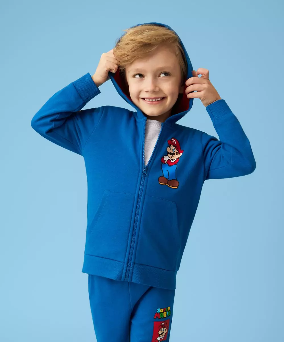 Chłopiec w niebieskim dresie z nadrukiem Super Mario, uśmiechający się z rękami przy kapturze.