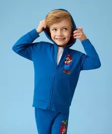 Chłopiec w niebieskim dresie z nadrukiem Super Mario, uśmiechający się z rękami przy kapturze.