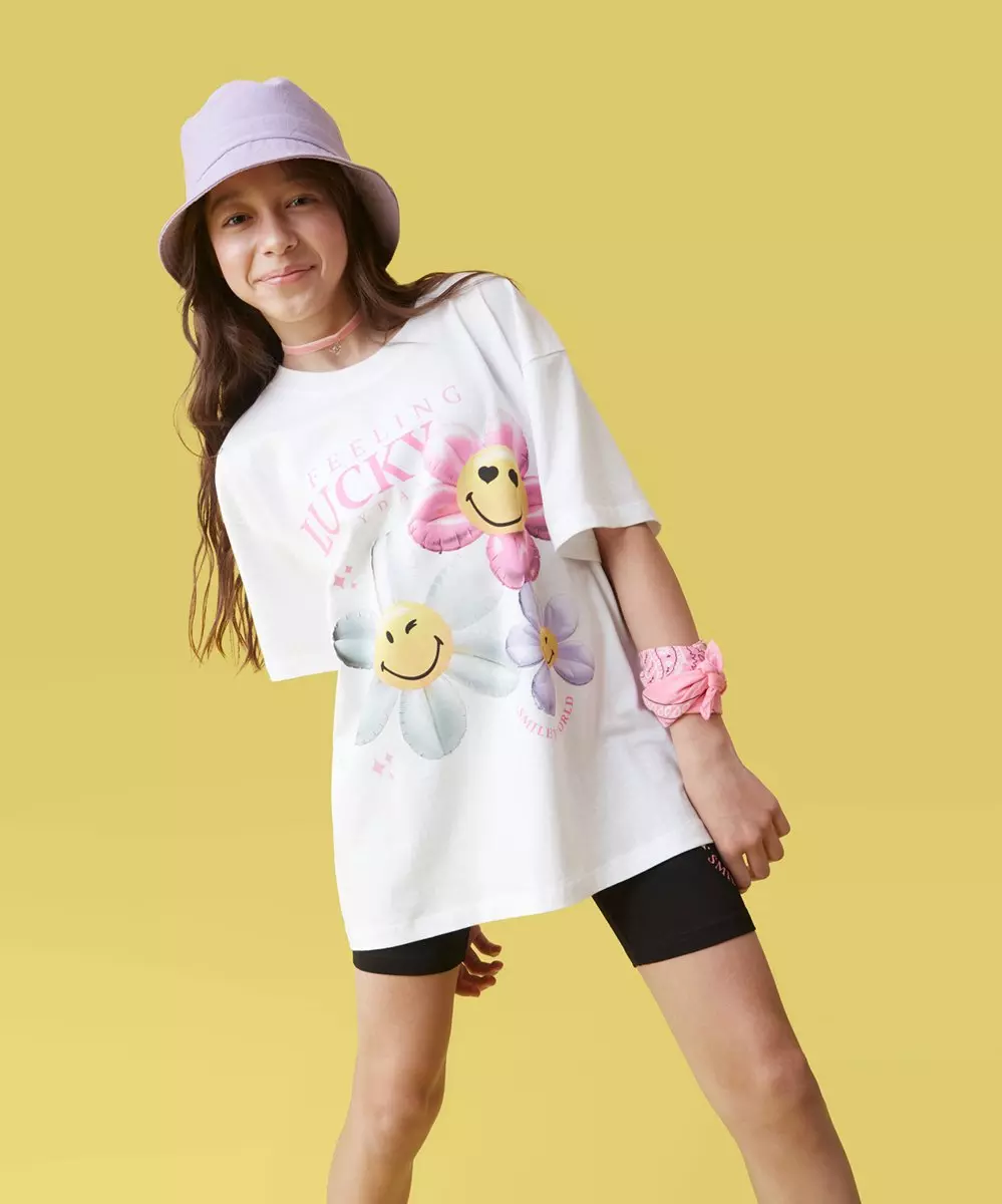 Fată purtând o pălărie mov și un tricou alb supradimensionat cu flori zâmbitoare și textul FEELING LUCKY.