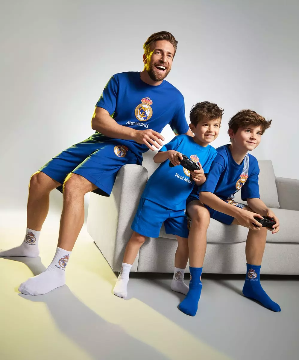 Tată și doi băieți purtând echipamente de fotbal Real Madrid albastre de la Pepco, jucând jocuri video pe canapea.