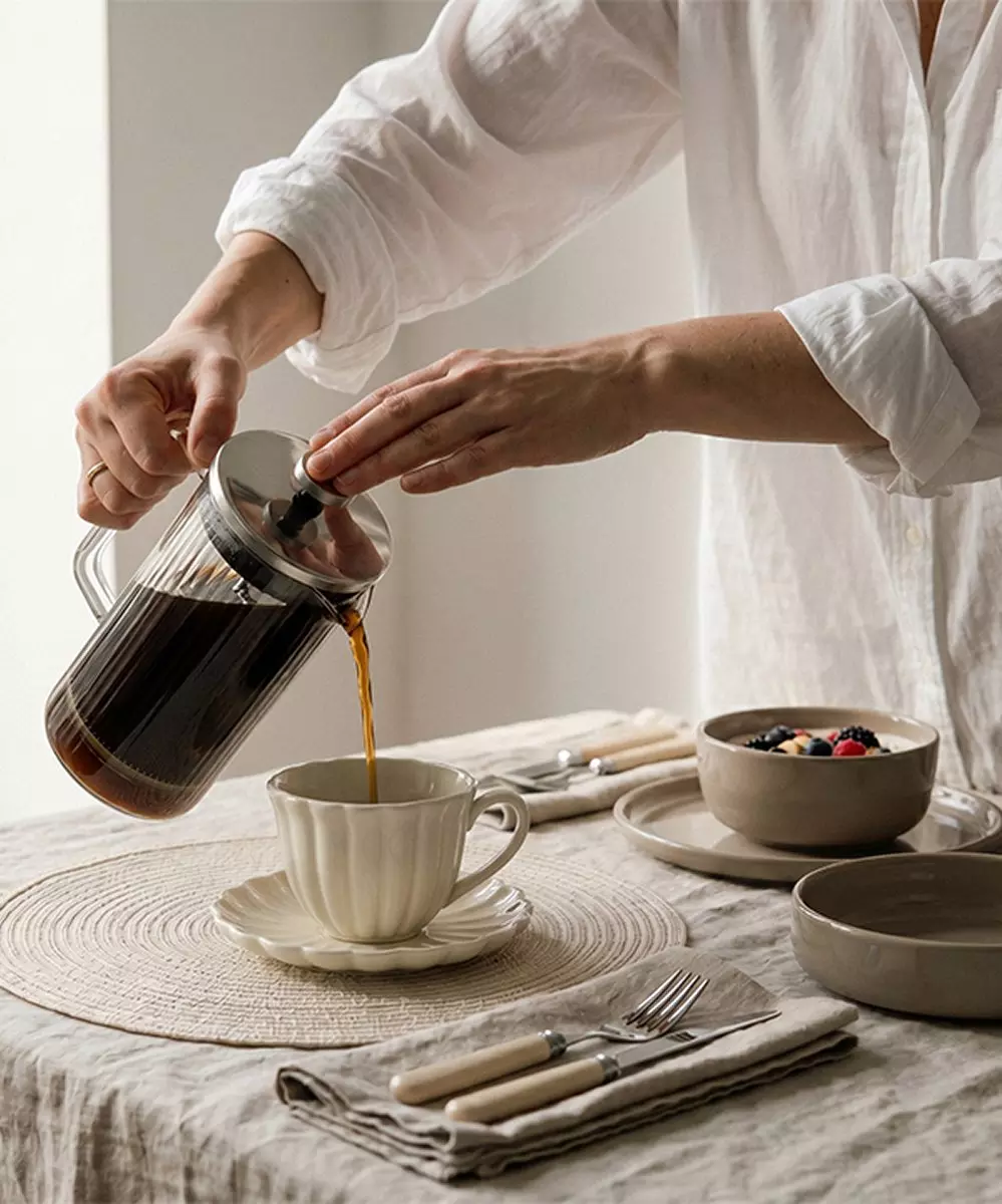 Persona sirviendo café de una cafetera de prensa francesa Pepco en una taza sobre una mesa de desayuno decorada.