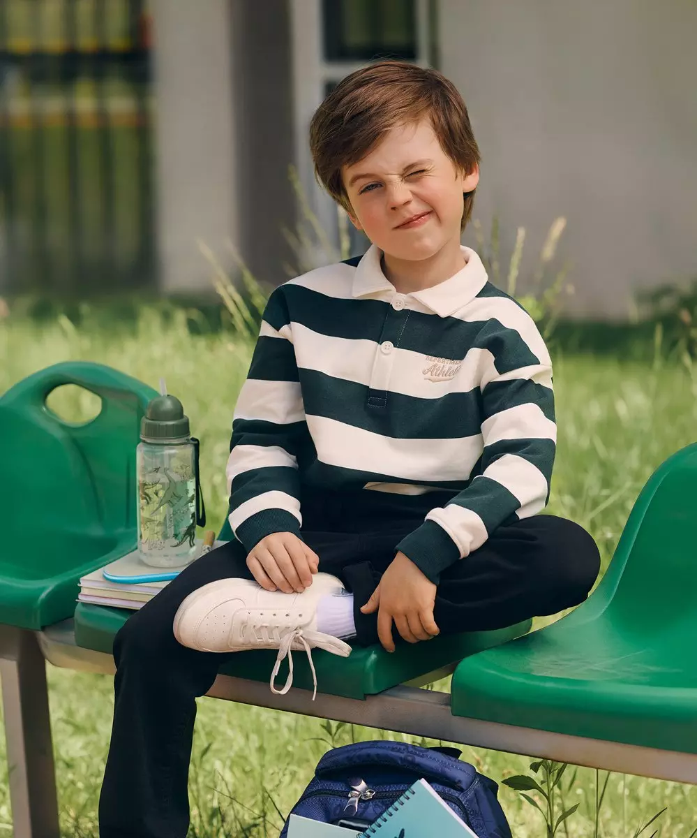 Băiat purtând tricou polo cu dungi Pepco, așezat pe bancă cu rucsac și sticlă de apă lângă el.