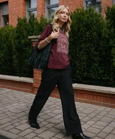 Woman walking outdoors wearing Pepco burgundy embroidered top with black trousers and fringed bag.