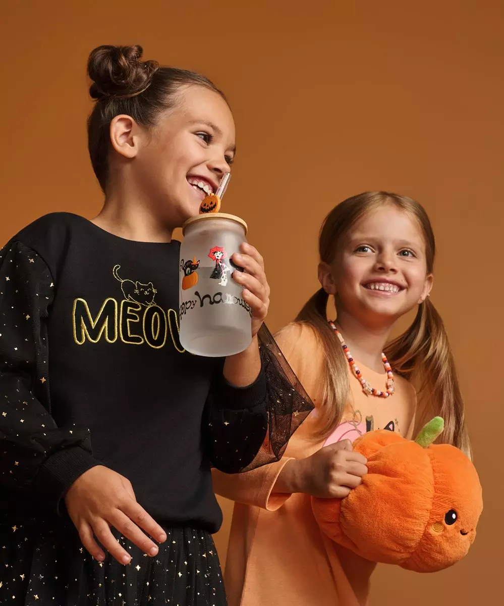 Dve dievčatá v halloweenskych kostýmoch Pepco s tekvicovou taškou a hrnčekom na cukríky sa smejú.